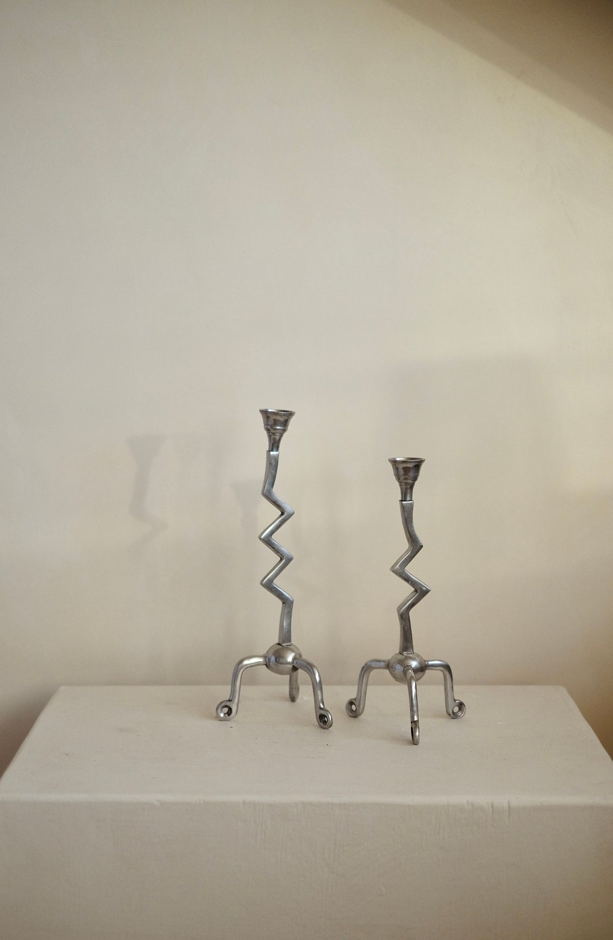 A pair of zig-zag shaped candle holders from septembre studios rests elegantly on small pedestals against a plain wall. Each features three outstretched feet, showcasing a modern, geometric design. Crafted in silver, the Set of Zigzag Candleholders is indeed a unique treasure for any collector.
