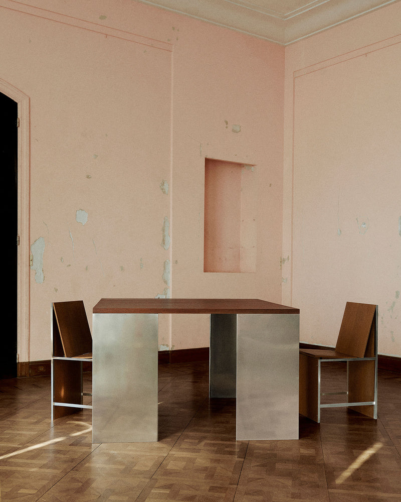 A minimalist room with peeling pink walls exudes grounded elegance, featuring Studio HAOS’s Square Table 2022—a modern piece with a wooden top and metallic legs—flanked by matching metal and wood chairs on a parquet floor.