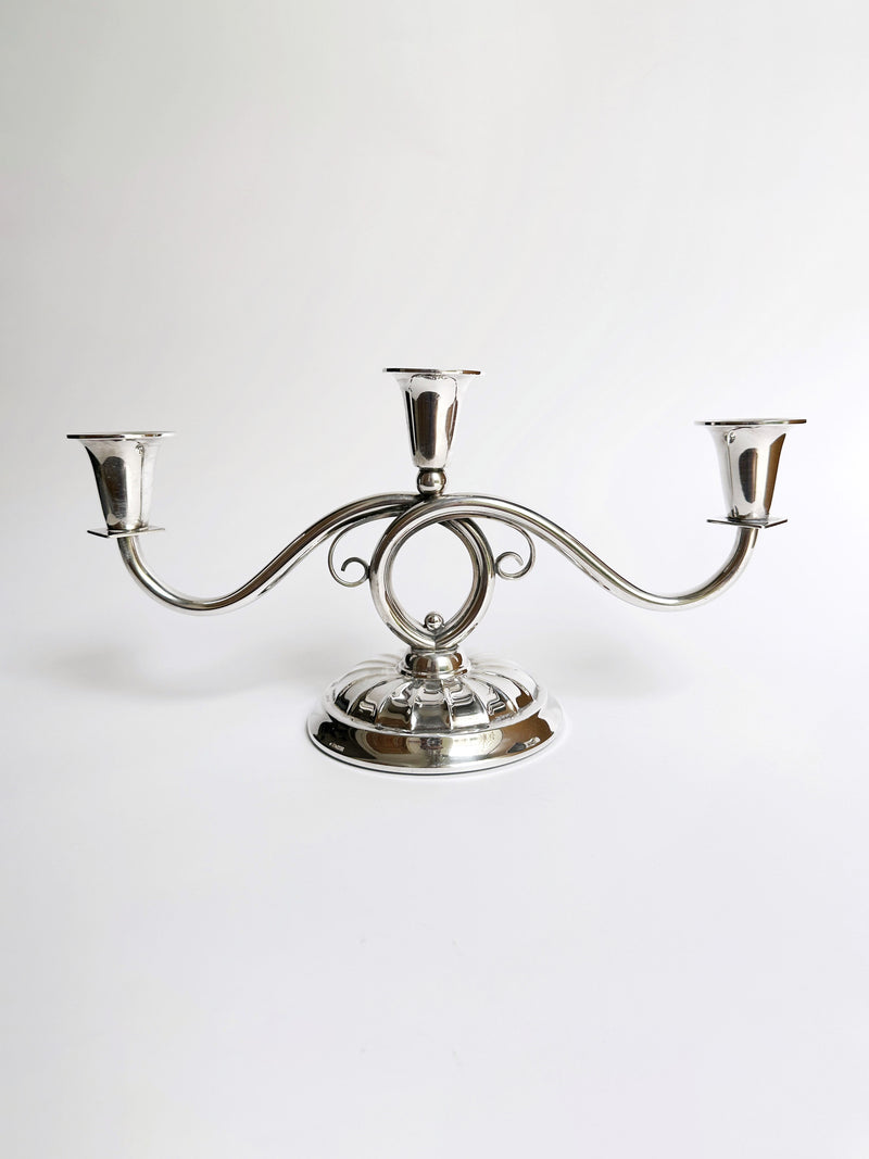 A Danish Mid Century Candleholder by Dodo Vintage, crafted in silver with an elegant, symmetrical three-arm design. The center arm boasts a circular loop, and the rounded base is adorned with decorative filigree details. This exquisite piece is showcased against a plain white background.