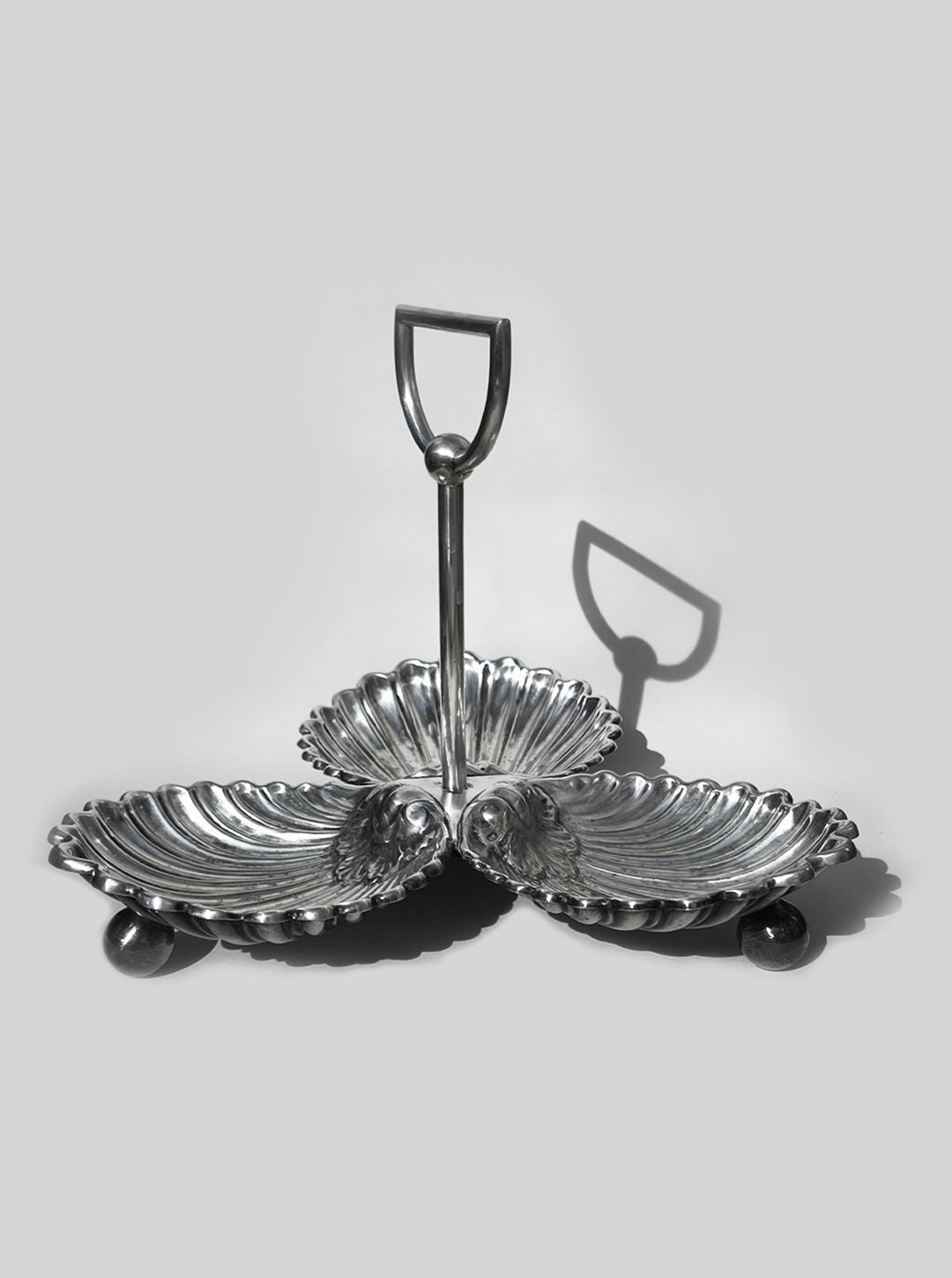 The Les Objoies Large Silver-plated Triple Tray features shell-shaped compartments forming a clover pattern with a central handle, casting an elegant shadow on a gray background—an eye-catching serving piece.