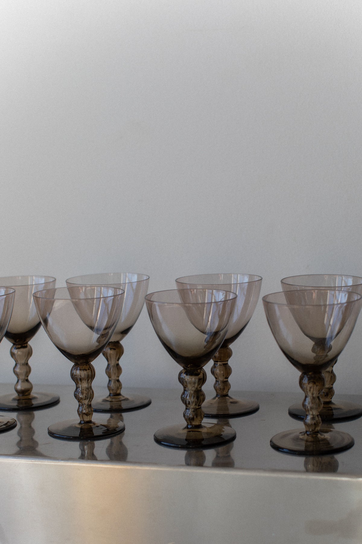 A row of elegant, Art Deco Glasses by Out For Lunch, featuring a vintage-style design with a smoky tint, crafted for a sophisticated drinking experience. The glasses are displayed on a reflective surface, showcasing their intricate and ornate stems as they are arranged neatly in a symmetrical manner against a plain background.