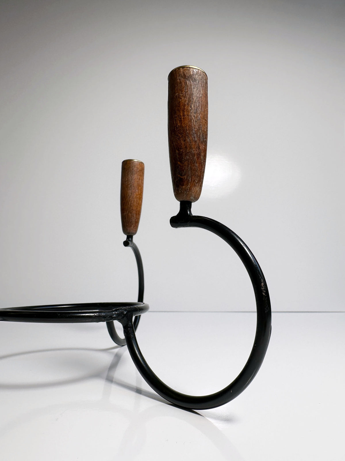 A minimalist photo showcasing two circular black metal rings with teak ends elegantly positioned against a plain gray background. The rings' clean lines and handles' natural wood grains highlight the simple yet stylish design of the Danish Candleholder 1960s from Dodo Vintage.