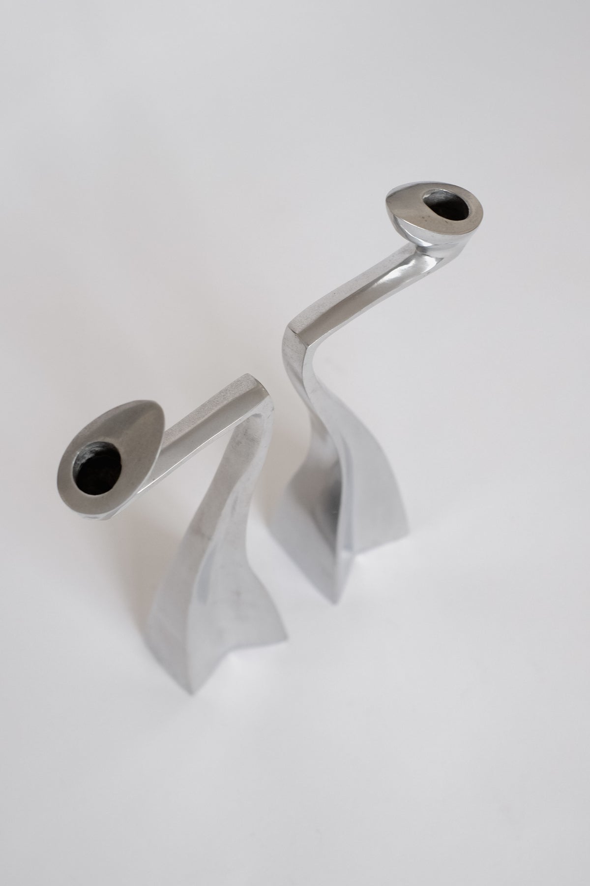 On a white surface stand two sleek, modern metal candle holders with curved designs from septembre studios. These Matthew Hilton creations from the '80s feature smooth, reflective finishes and abstract shapes, offering both minimalist appeal and artistic flair as collector's items.