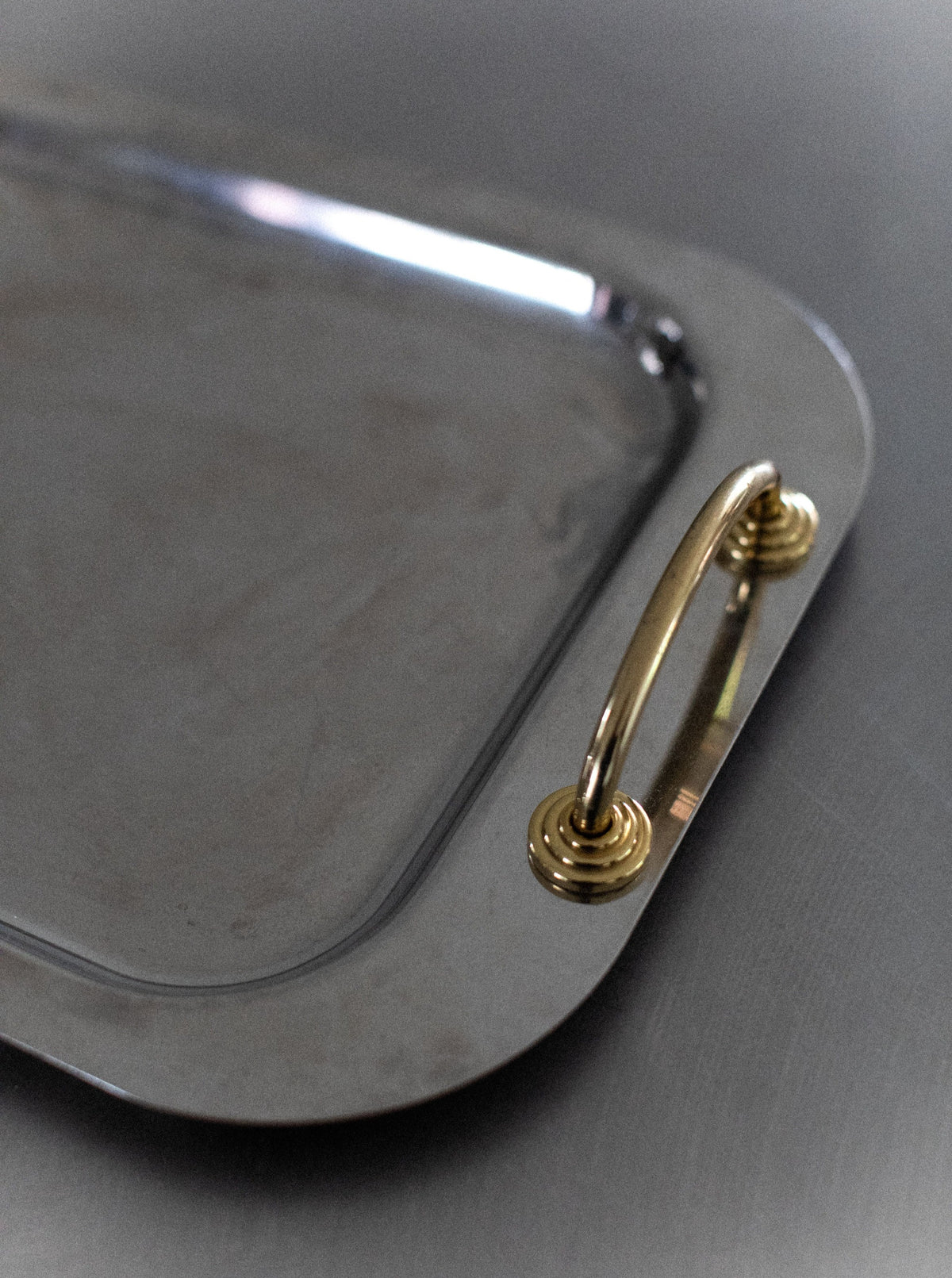 A close-up image of a Stainless Steel Tray with a shiny surface, featuring a single brass handle on the right side. The handle is attached to the tray with two screws, and the tray's edges are slightly rounded. The modern elegance of this design complements the metallic background surface. This exquisite piece is by Out For Lunch.