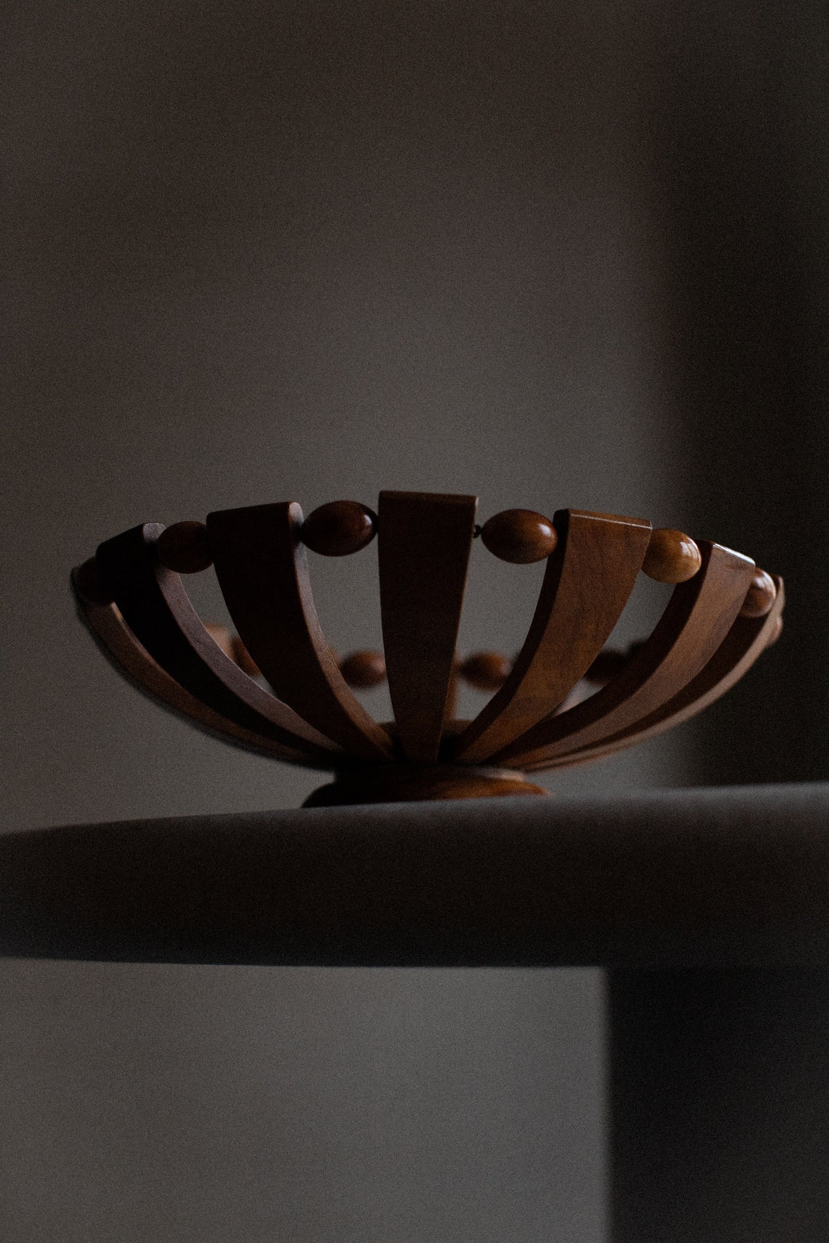 The Out For Lunch 1950's Wooden Bowl features open, curved slats and round accents that display natural grain, adding rustic charm as decor on a light tabletop against a dark, blurred backdrop.