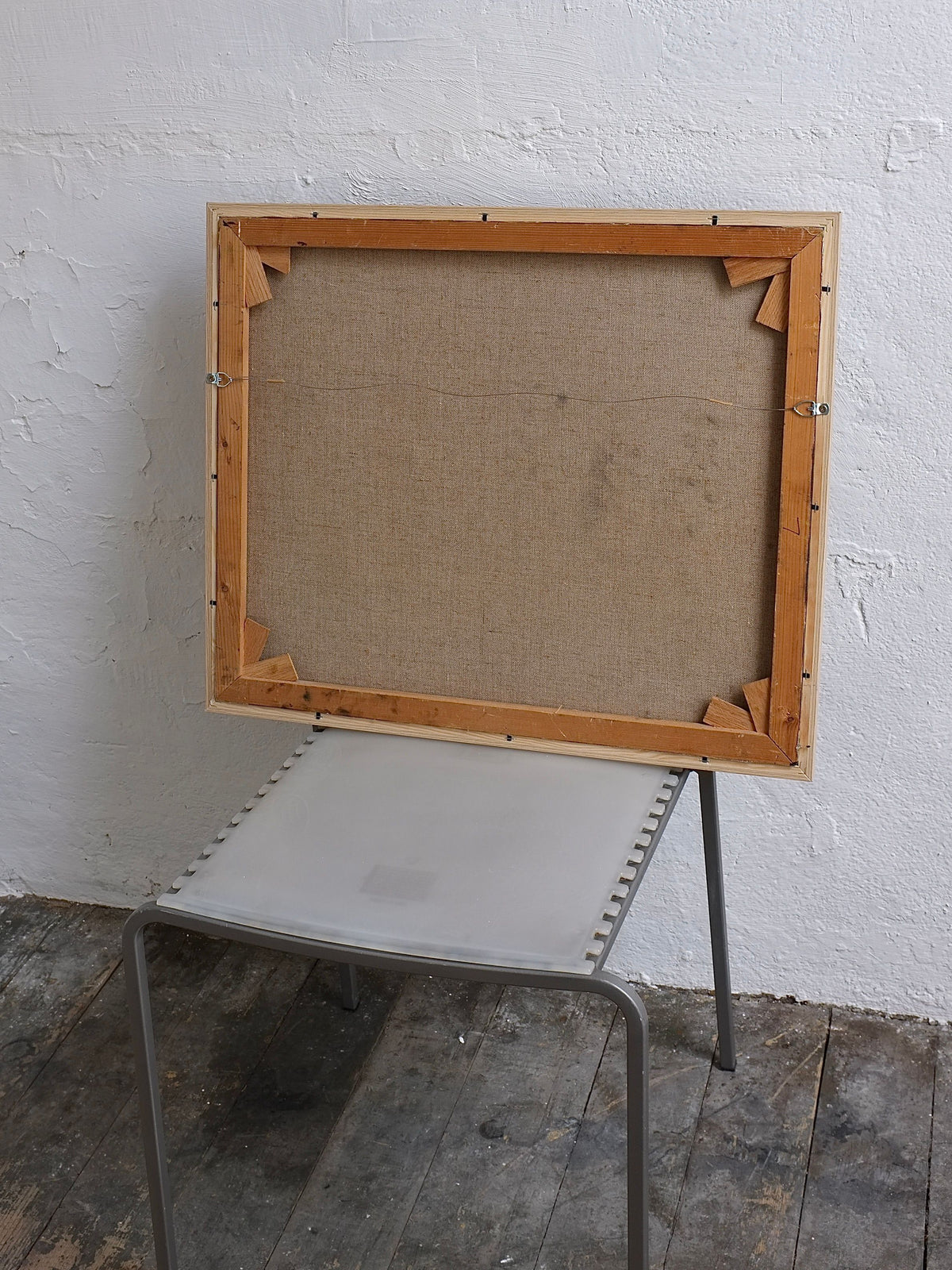 A Veter Vintage Oil on Canvas Landscape Painting with earthy tones rests on a simple metal chair, its wooden frame against a textured white wall, while worn rustic wood planks underfoot evoke the timeless charm of the Netherlands.