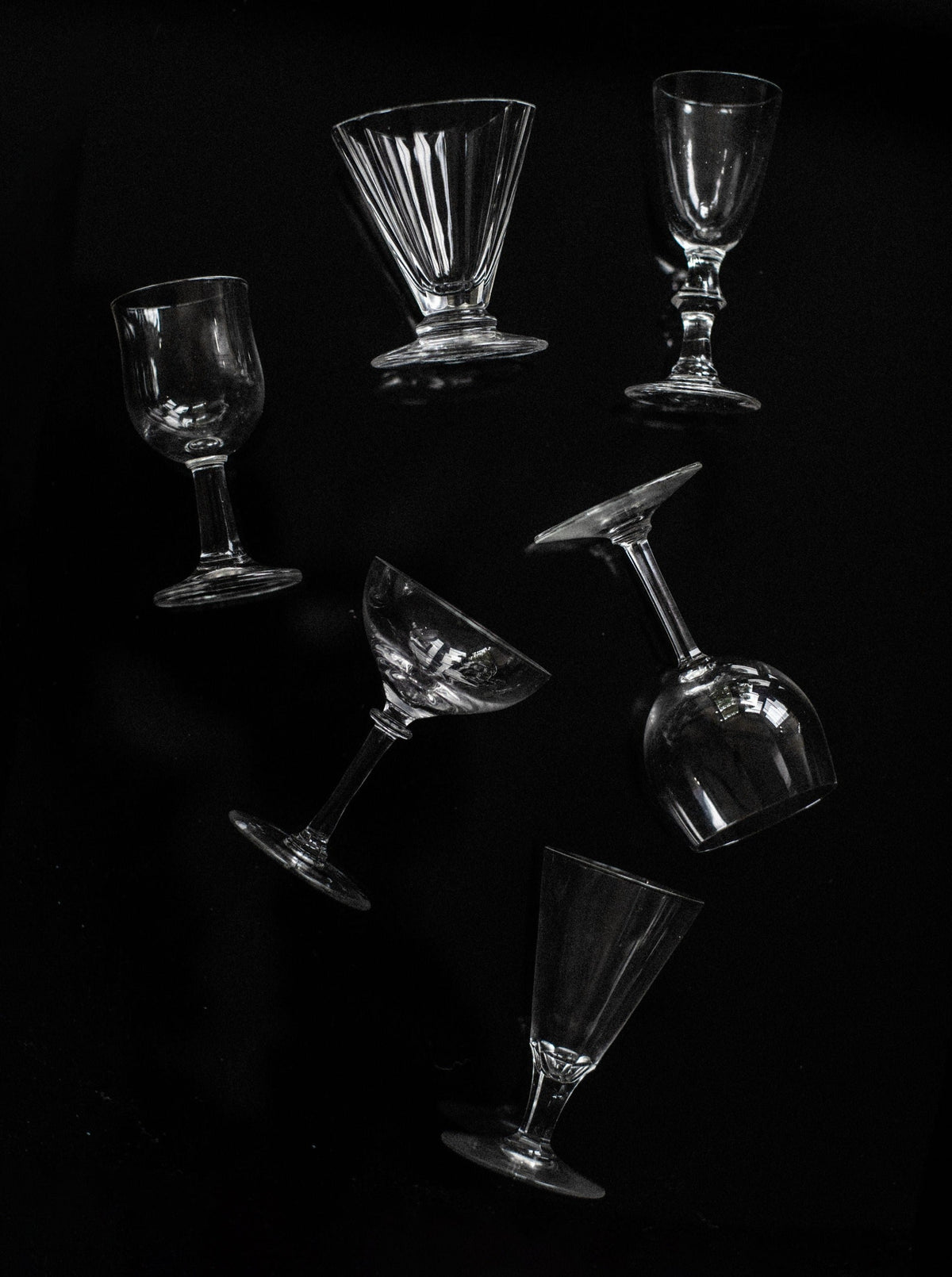 A set of six mix-matched glasses by Out For Lunch, featuring diverse types of empty glassware such as wine glasses, cocktail glasses, and goblets, are arranged diagonally against a dark background. Each curated piece is oriented in various directions, creating an artistic and dynamic composition for a sophisticated drinking experience.