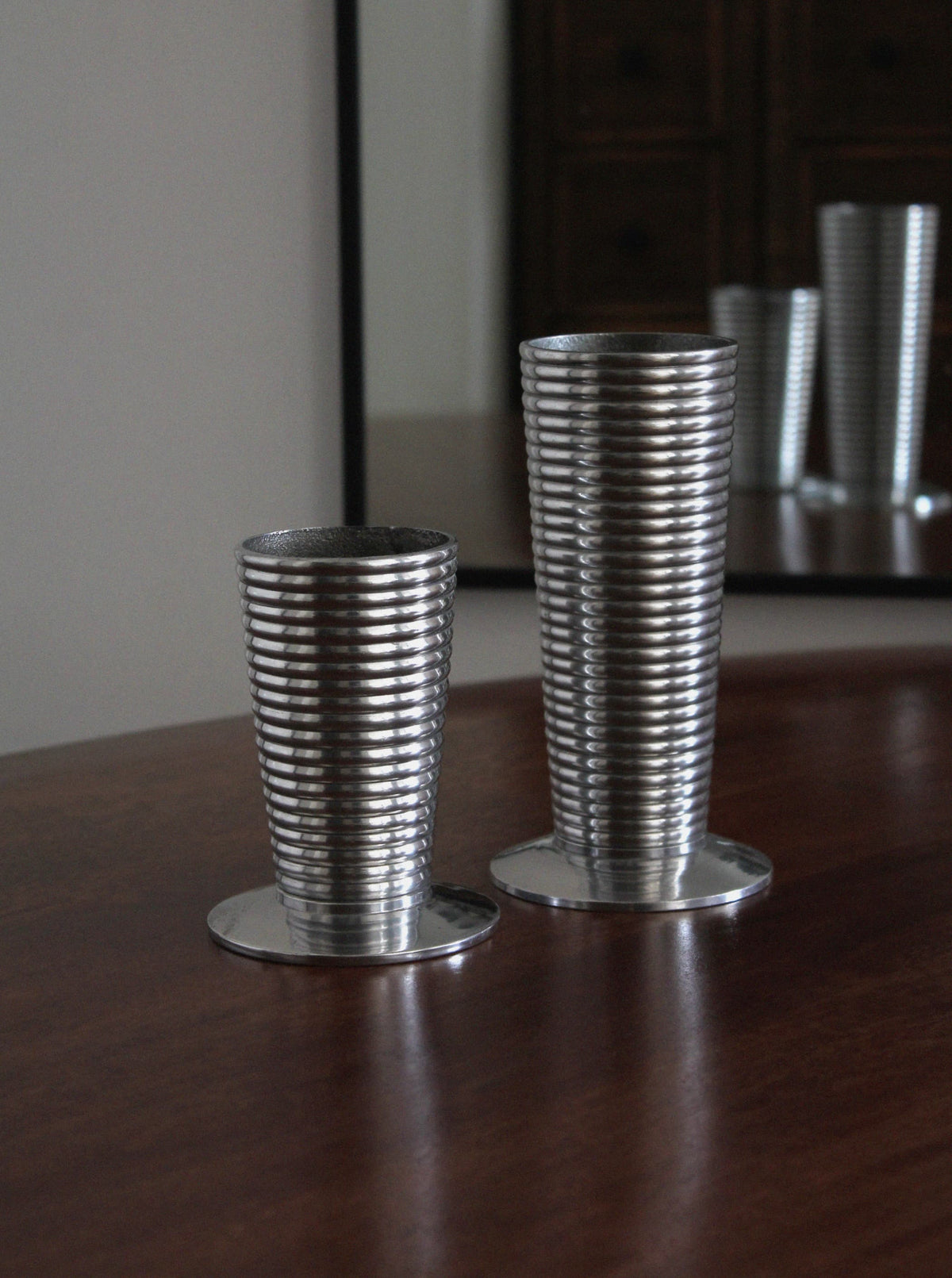 The Pair of Aluminum Vases by Vintage Stories, featuring sleek silver ribbed designs in two heights, stand on a wooden surface and reflect modern elegance in the mirror behind them.