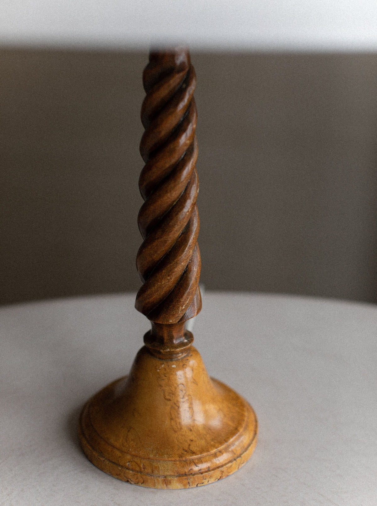 A close-up view of an Out For Lunch Twisted Wooden Lamp base resting on a light-colored circular surface. The polished finish highlights the intricate spiral design and natural wood grain, showcasing exquisite artisanal craftsmanship. The background is plain and slightly out of focus, emphasizing the lamp's beauty.