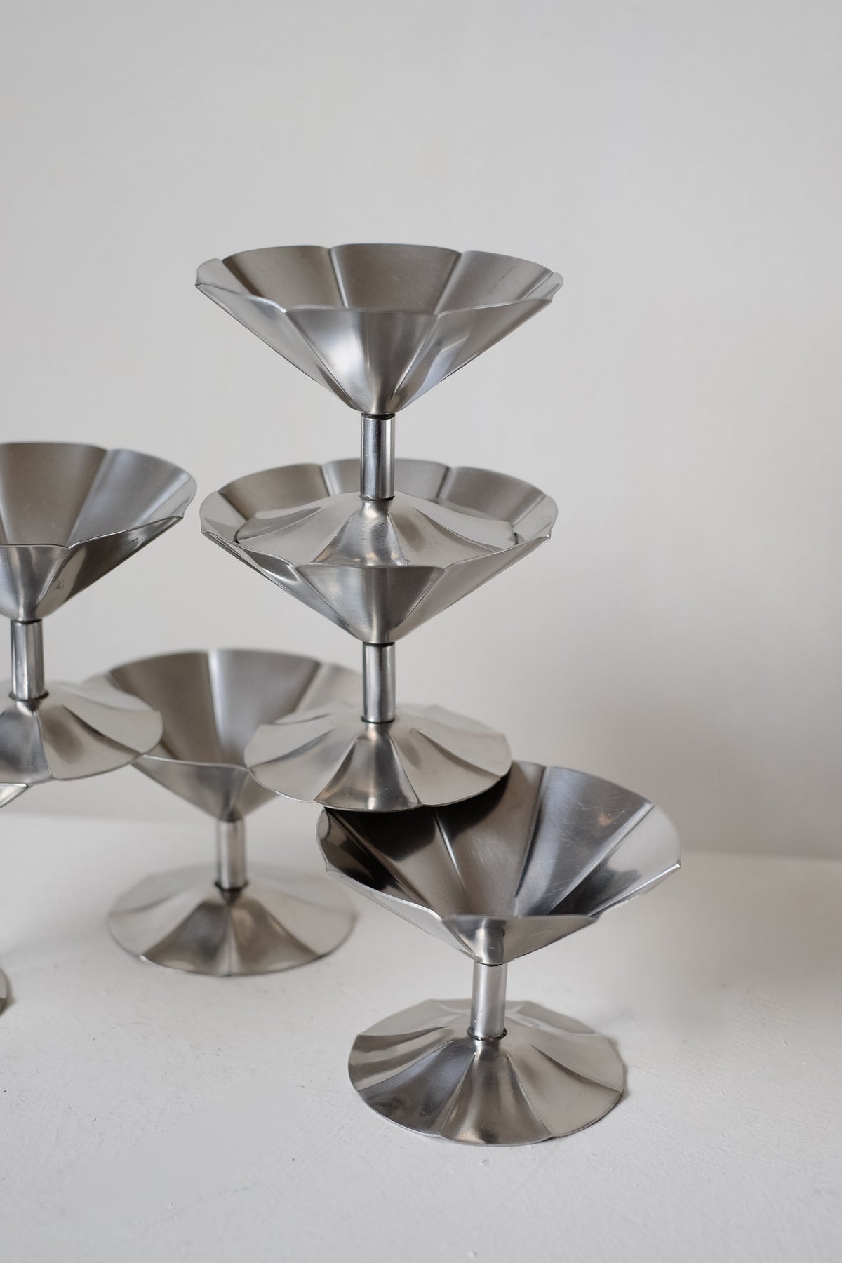 A pyramid of stacked shiny stainless steel cocktail glasses, part of the septembre studios' Set of 6 Ice Coupes, reflects light for a sleek, modern look against an off-white background.