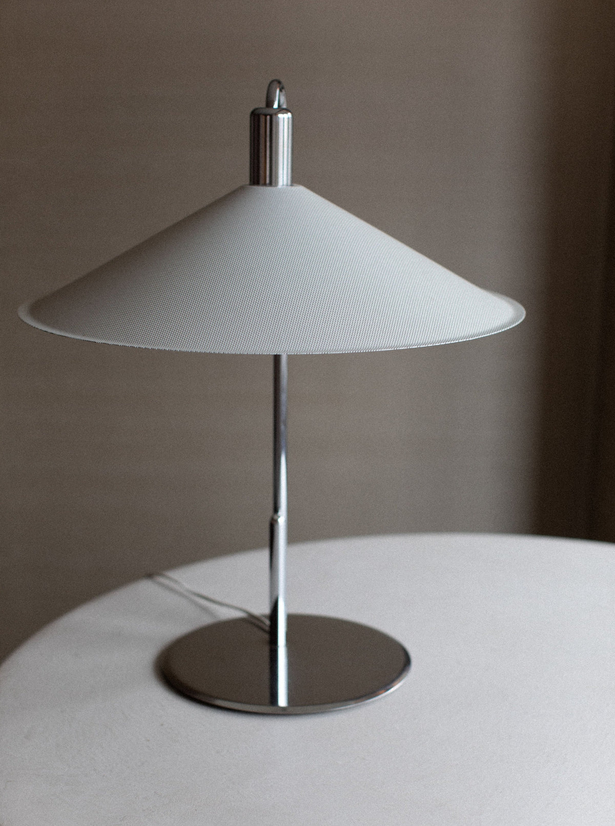 An Out For Lunch Börge Lindau Lamp with a conical white shade and chrome base sits on a white circular table against a neutral background. The lamp is turned off, showcasing its sleek and minimalist Scandinavian design.