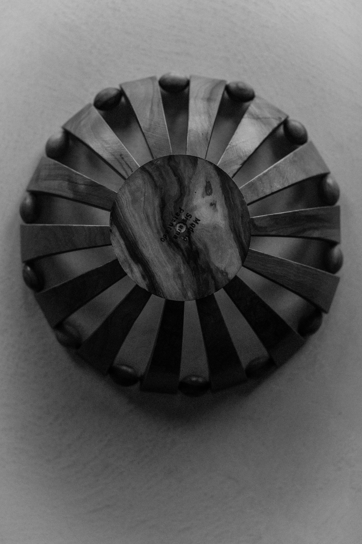 A black and white photo features the 1950's Wooden Bowl by Out For Lunch—a circular piece with a central wood disc and radiating curved metal segments with knobbed ends, mounted on a textured wall.