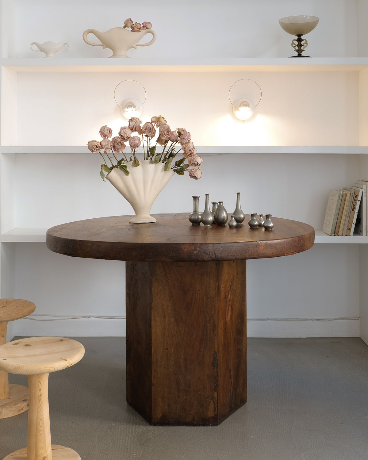 The Midcentury Hexagonal Wood Dining Table by Collection Apart showcases a large beige vase with wilted flowers and several small metal vases. Behind it, white shelves filled with decorative items and books set the scene, as two wooden stools peek into view.
