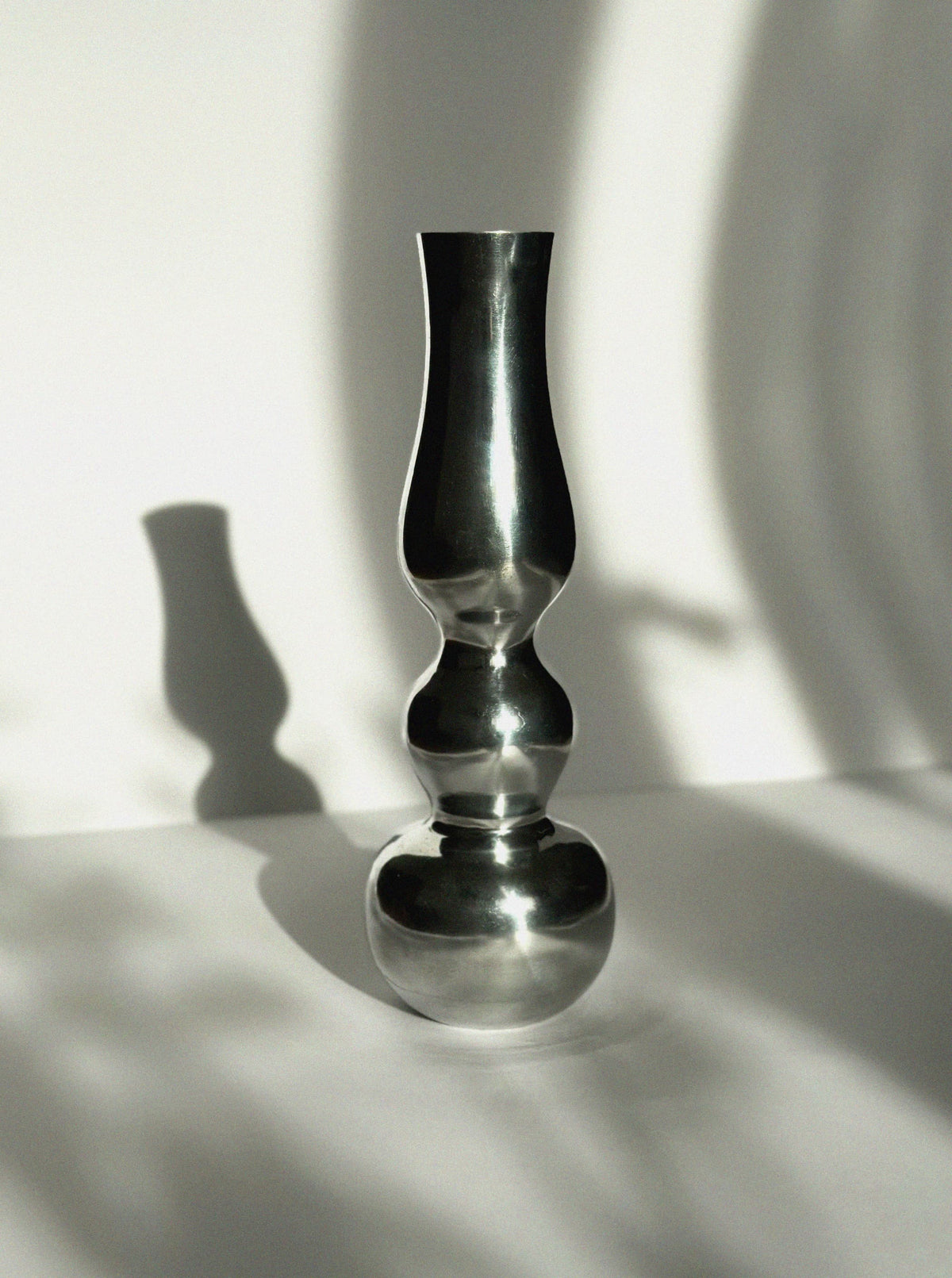 The Aluminum Figure Vase by Vintage Stories features a tall, metallic form with a mirror finish and sculptural curves. Set on a white surface, it casts bold shadows while blurred branches in the background highlight its minimalist elegance.