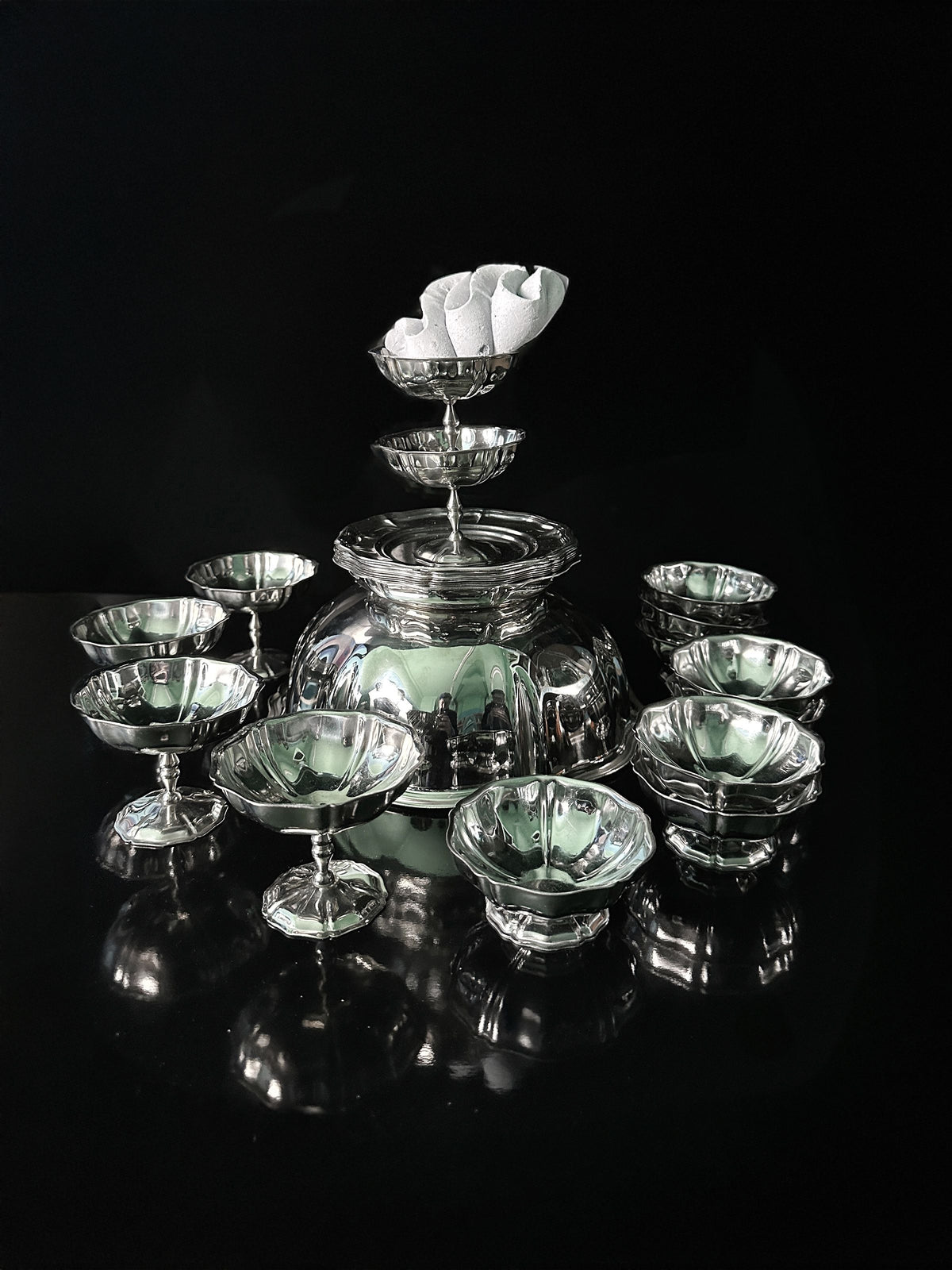 Reflecting light against a black background, the Dodo Vintage Italian Stainless Steel Dessert Coupe Set features an artistic arrangement of multiple small cups surrounding a central bowl, with one cup elegantly placed on top.