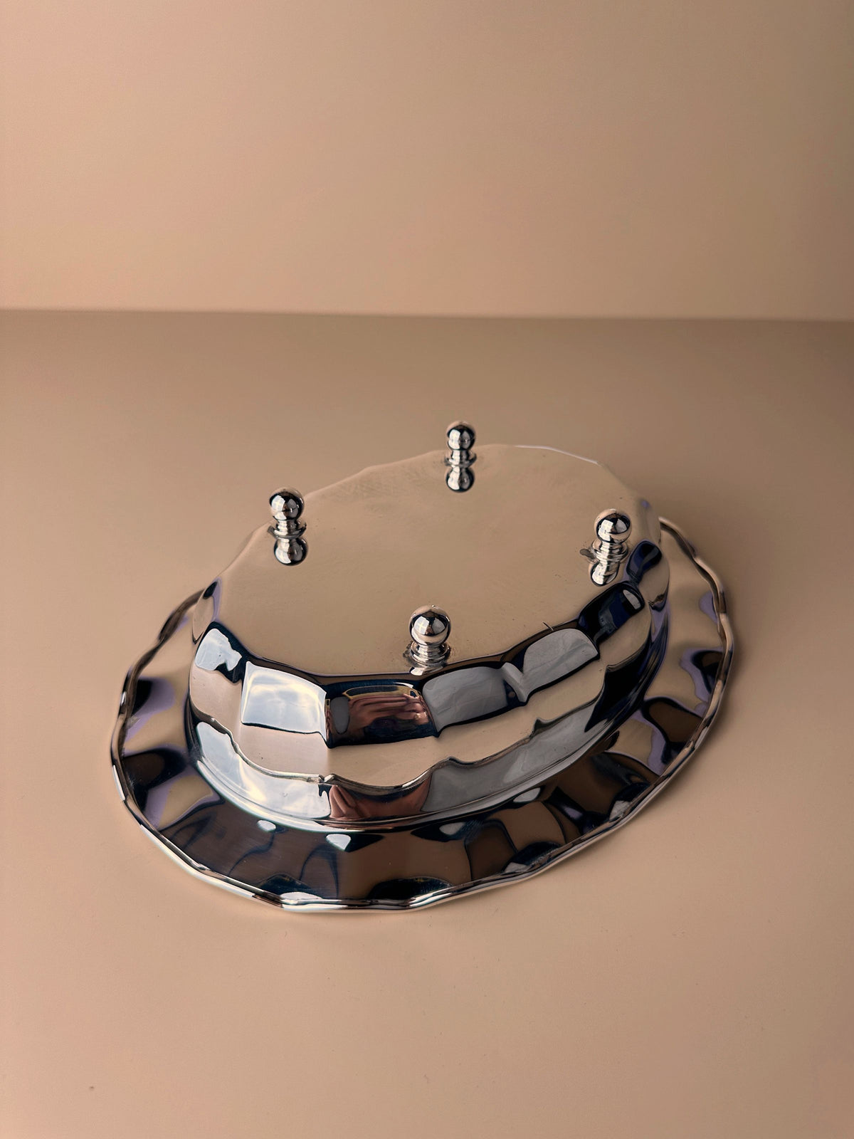 A reflective, silver-plated Italian footed bowl with an irregular oval shape and rounded edges sits on a flat beige surface. Three small metallic pegs protrude from the top. The "Mid Century silver plated italian bowl" by Dodo Vintage and its surroundings exhibit a minimalistic aesthetic with soft lighting.