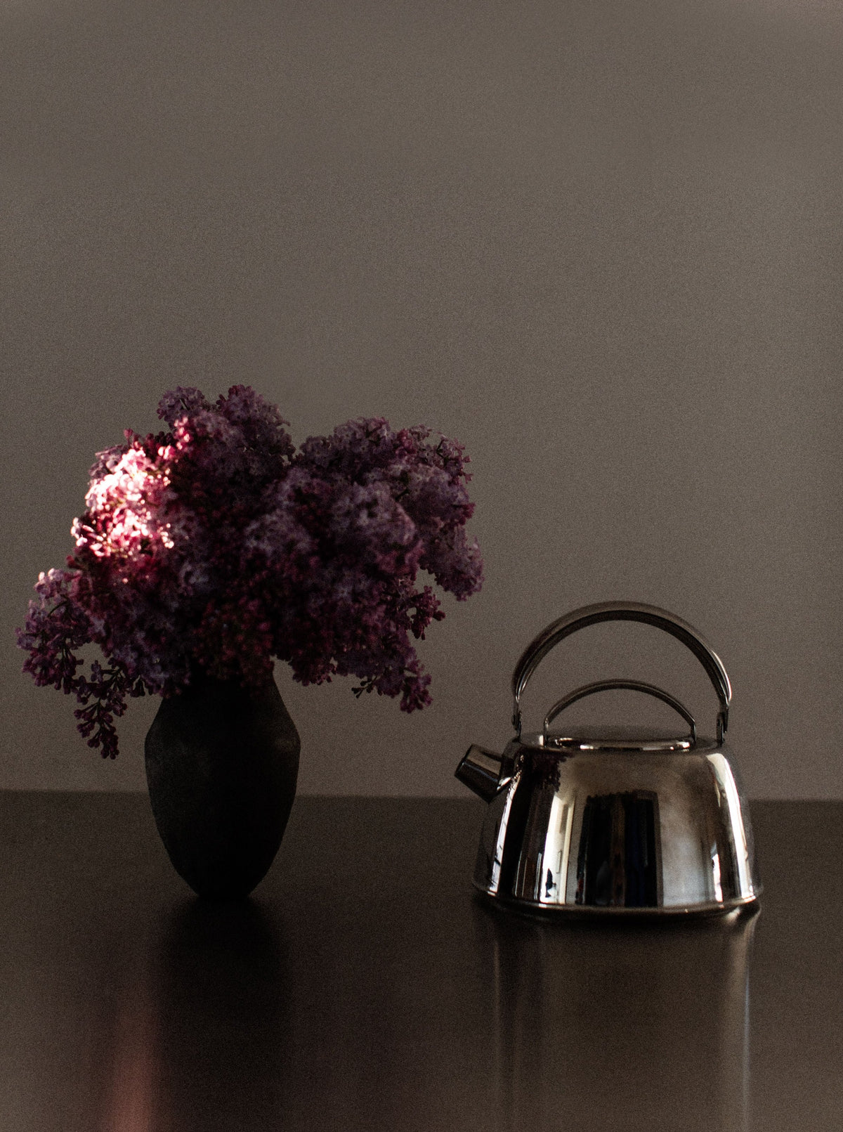A dimly lit scene features a dark vase filled with vibrant purple flowers on the left, and an Out For Lunch Chrome Teapot on the right, both placed on a reflective dark surface against a plain brownish background, enhancing the overall aesthetic appeal.