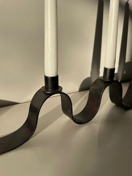 Close-up of The Ribbon Candelabra by Maud Vaughan, a handmade black metal piece with a wavy base, holding white taper candles. Sunlight casts soft shadows on the white surface and wall behind.