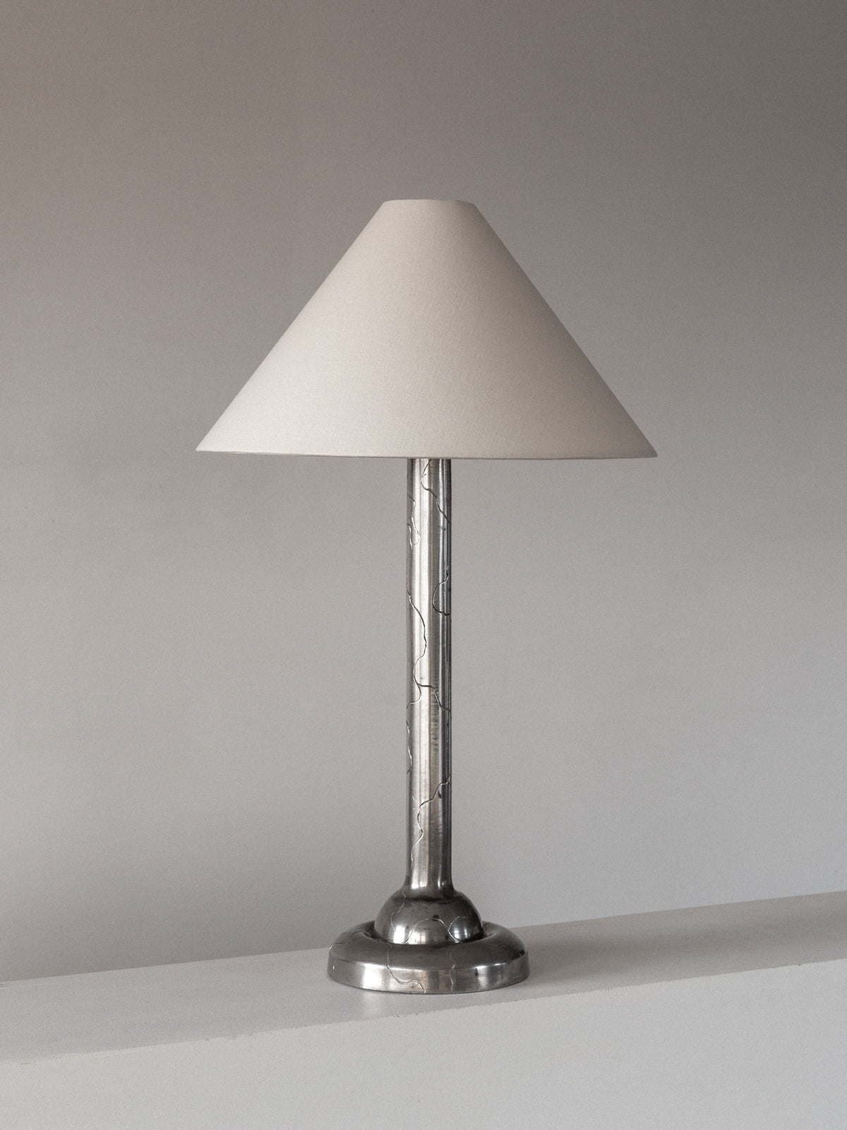 The Spigel Art Deco Pewter Lamp from 1920s Sweden features a cone-shaped white shade and metallic base, set on a white surface against a plain gray background.
