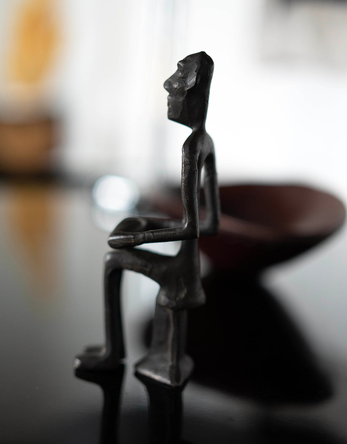 Porter Studio's Bronze Drummer Man Sculpture features a small, dark bronze seated figure with elongated limbs and minimalist details, delicately carved while holding a bowl as if drumming softly. Both items are set on a glossy surface against a blurred background.