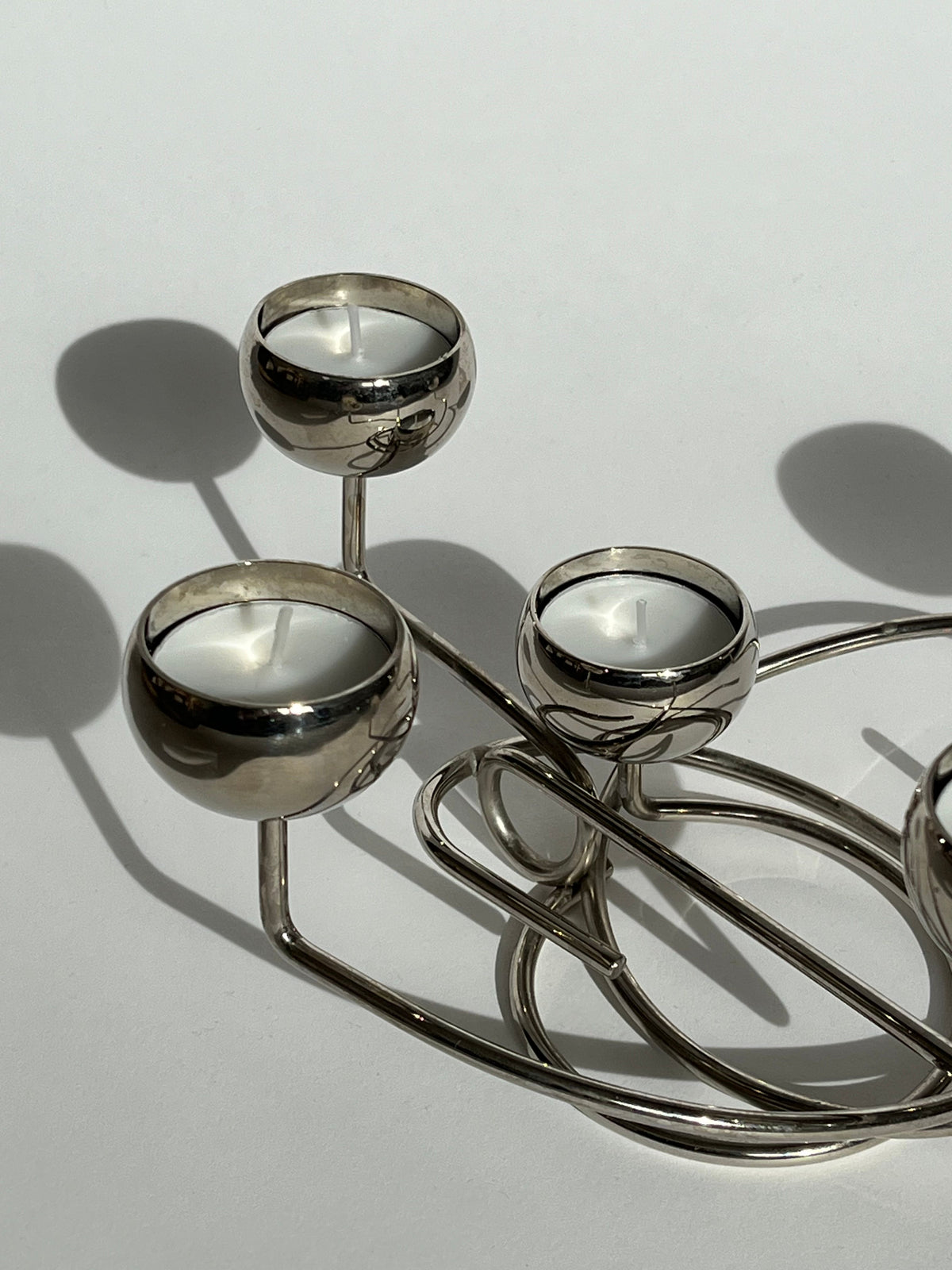 Introducing the Chrome Candleholder by septembre studios: a decorative metal piece featuring four round cups, each holding a small white candle. Its polished chrome finish and unique geometric design create soft shadows on any light background.