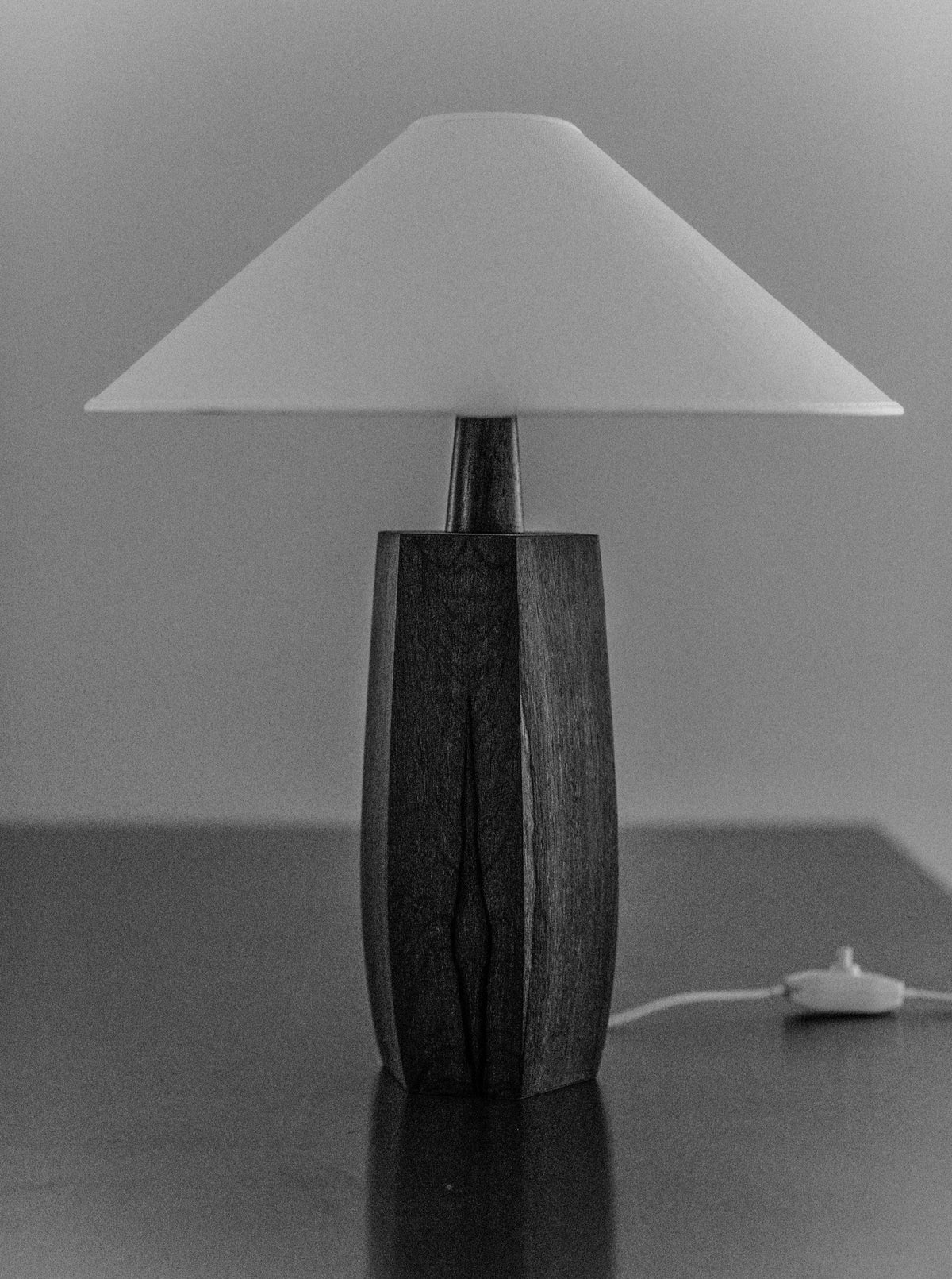 A black and white photo of a 1940's Wooden lamp by Out For Lunch with a textured wooden base, showcasing mid-century design, and a smooth, conical lampshade. The lamp is placed on a flat surface, and a cord with a switch is visible beside it.