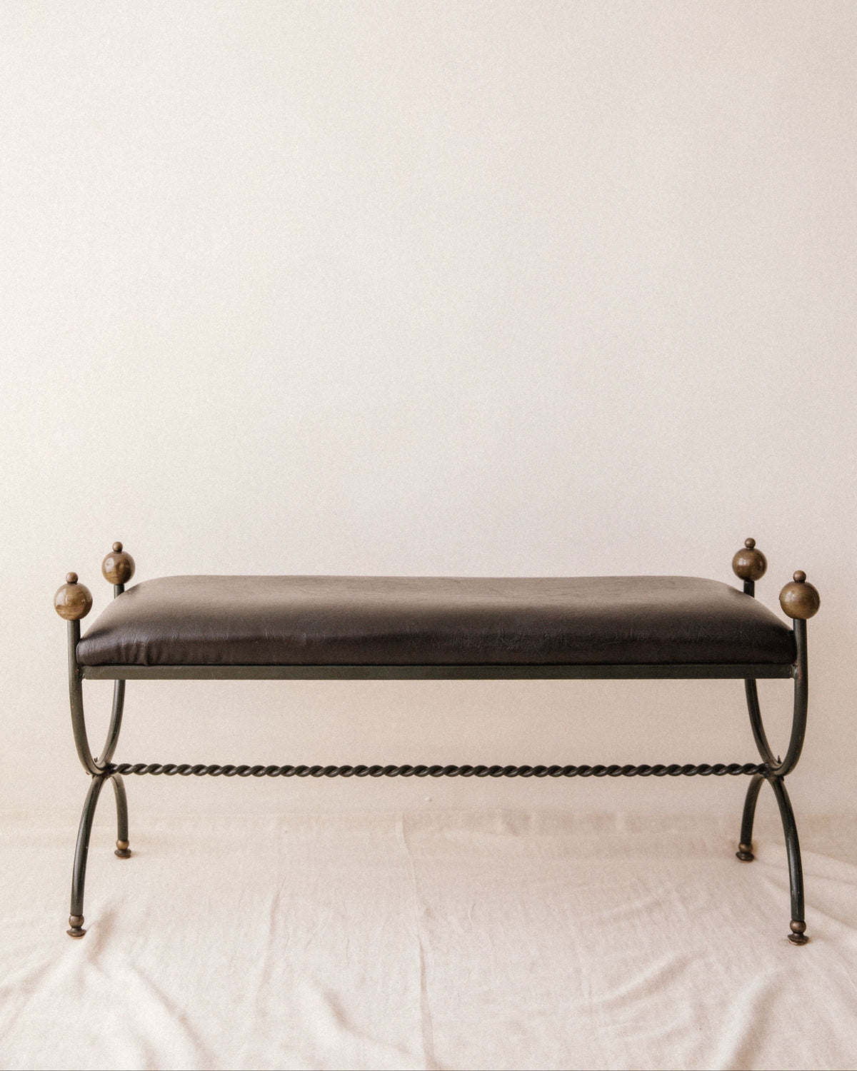 The Mid-Century Wrought Iron Bench by Arcā Collections features a dark cushioned seat, black metal frame, twisted legs, and round finials on each corner, evoking Jean Royère's style against a plain light backdrop.