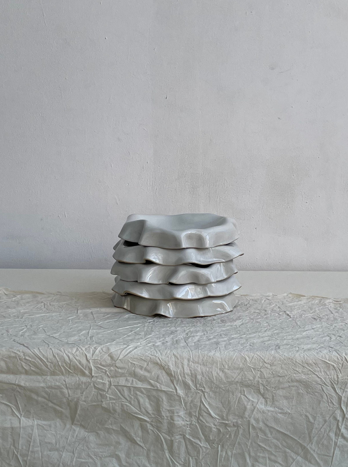 Set against a plain light grey wall, a stack of four Melted Butter Plates in Off White by LESS sits atop crumpled off-white fabric covering a flat surface. These irregularly shaped, wavy-edged plates with a glossy off-white glaze evoke the feel of handmade stoneware ceramics as minimalist, abstract art pieces.