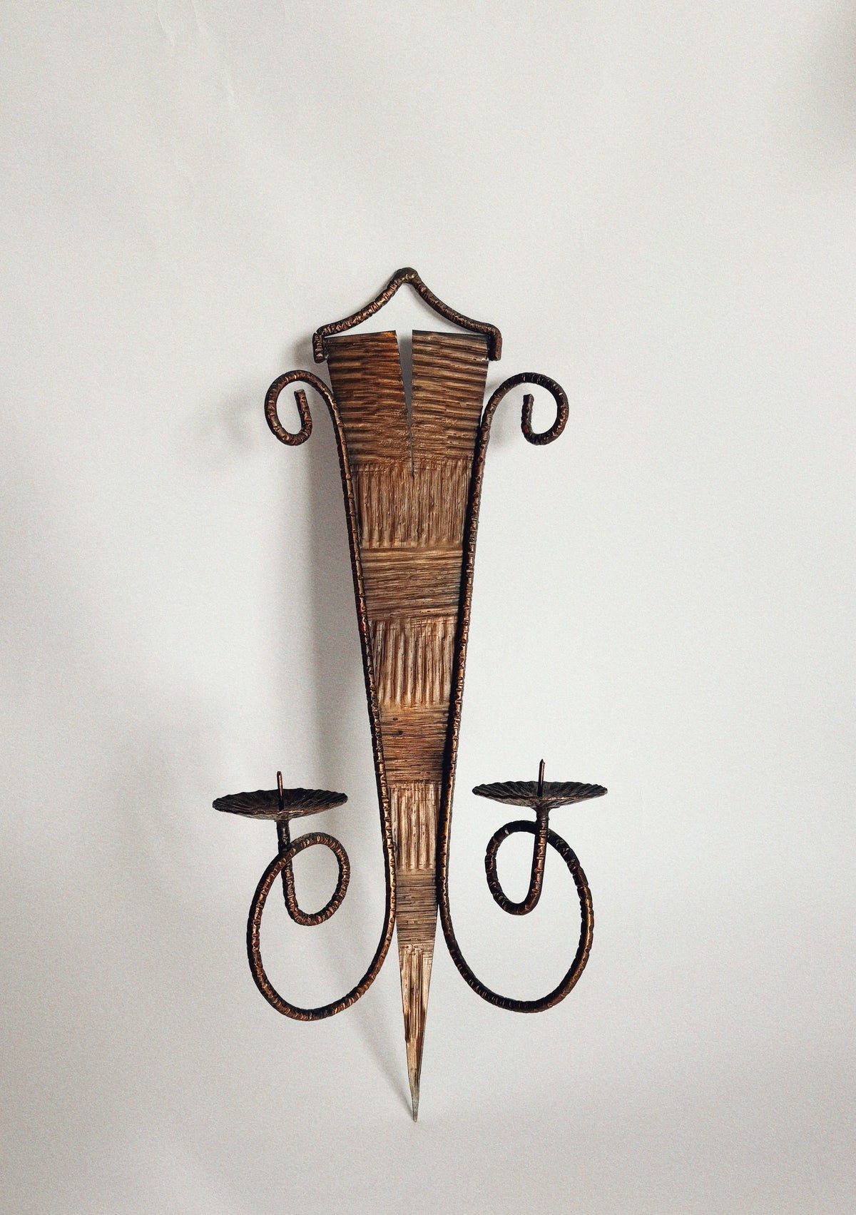 The Huerto Metalwork Wall Candleholder features a pointed, textured body, two curled arms with flat round holders, artisan design, and hammered details, displayed against a plain white background.