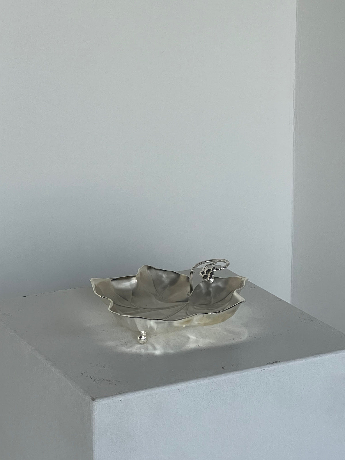 Displayed on a white square pedestal in a minimalist setting, the Grape Leaf Dish from Porter Studio is a vintage 1930s silver piece, shaped like a leaf with an elegant small handle and ball feet.