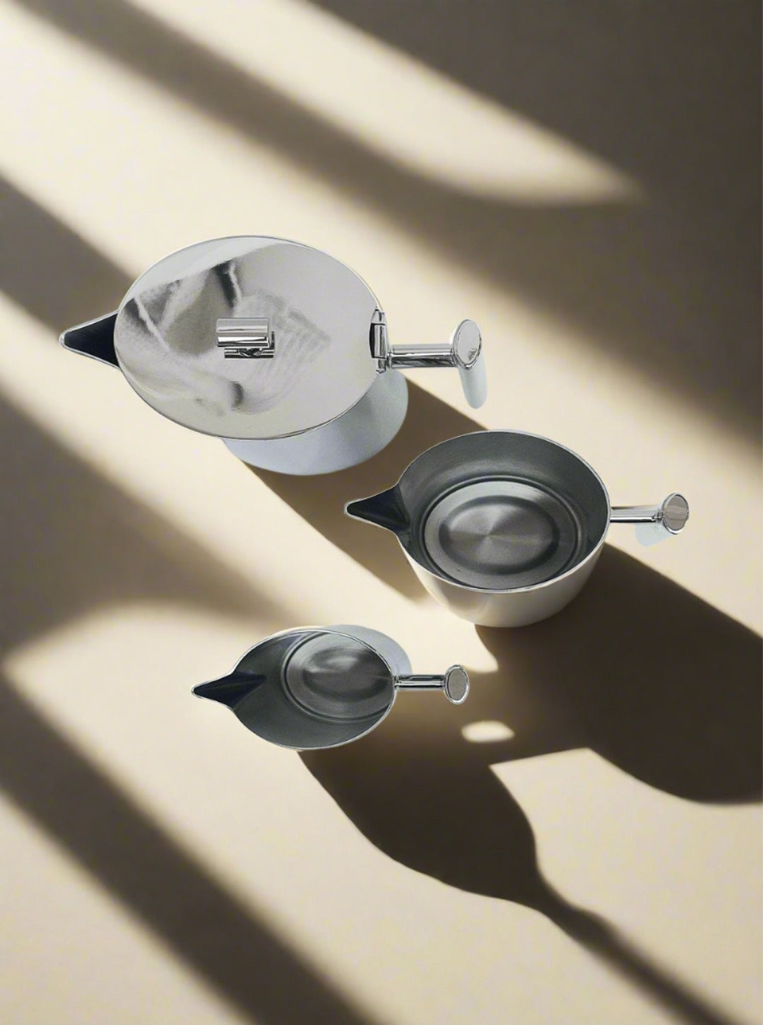 Three modern, minimalistic stainless steel teapots with sleek handles and spouts are arranged on a textured beige surface. The lighting casts dramatic shadows, highlighting their glossy finish and streamlined design, making them a perfect addition to any Les Objoies Vintage Coffee Set.