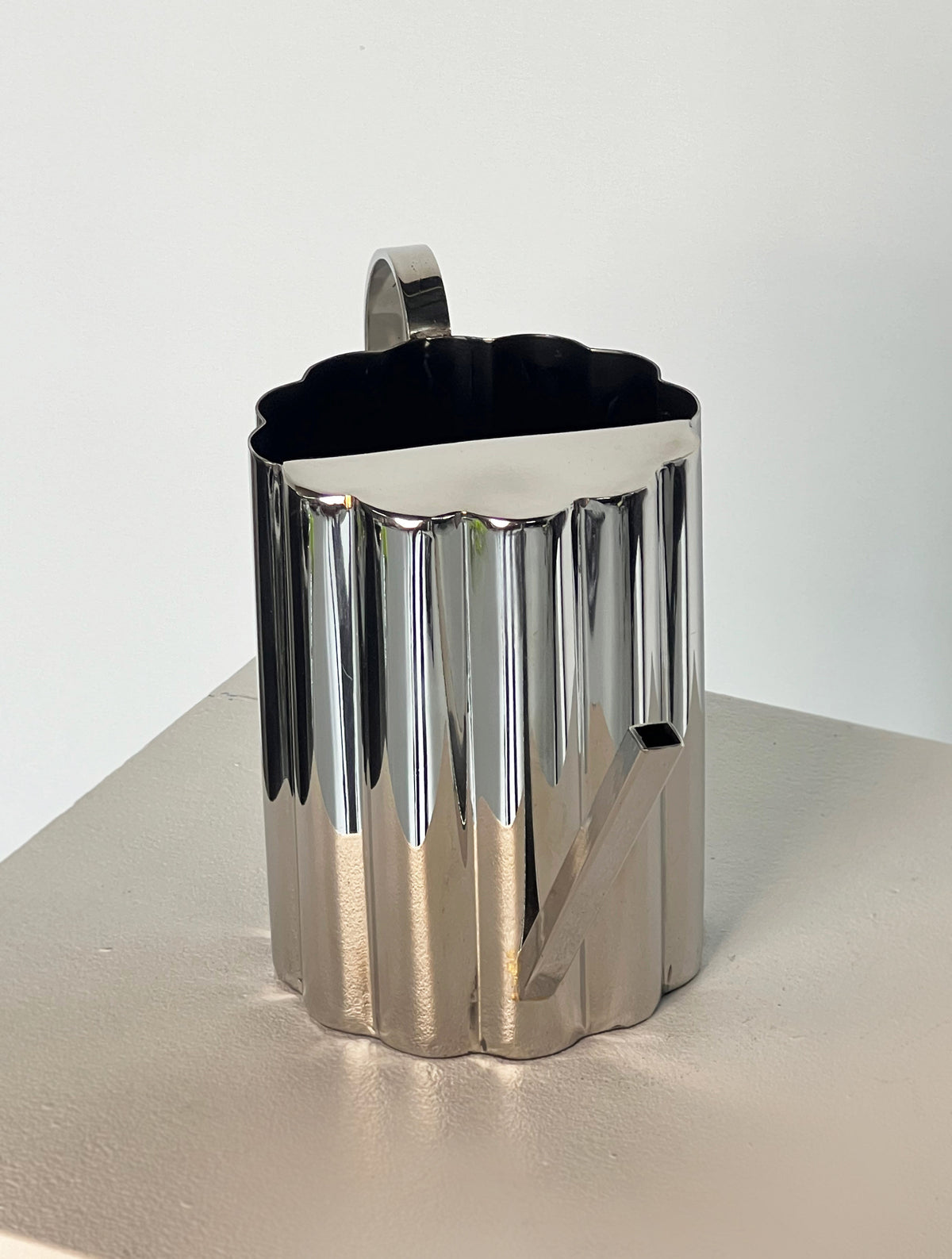 The Fluted Watering Can by Porter Studio features a metallic, scallop profile with a triangular spout and curved handle. It has a shiny, reflective finish and modern design, resting on a light-colored surface.