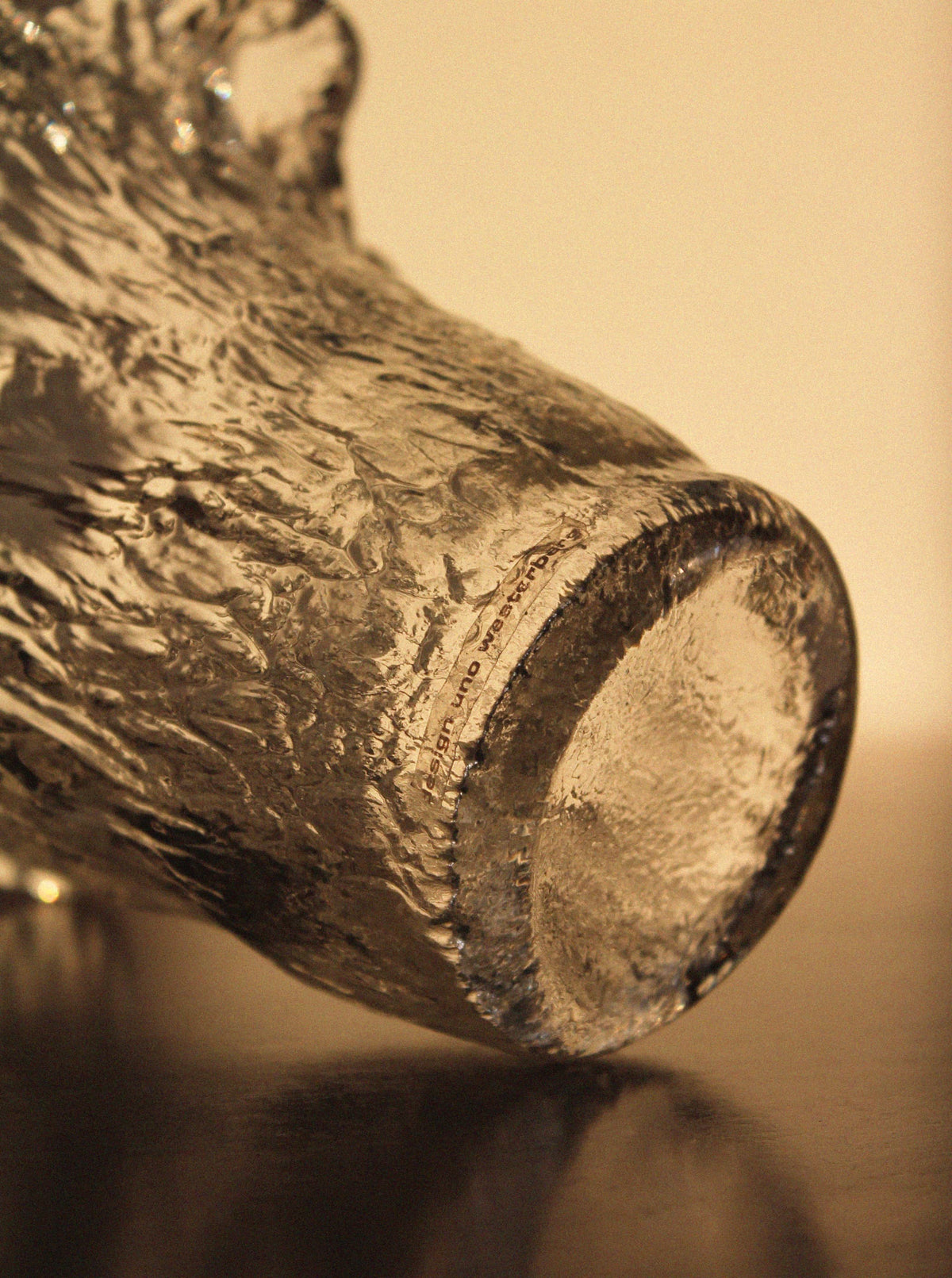 Close-up of the textured base of a Vintage Stories Uno Westerberg 60s Organic Glass Vase, clear with a rough, uneven finish, shown slightly tilted on a reflective surface.