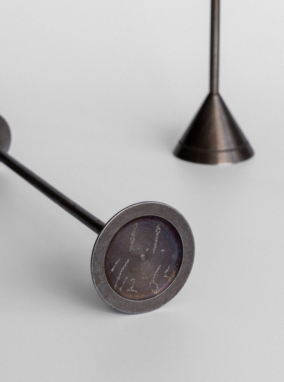 A close-up of two metal stamps with cylindrical handles showcases the detailed head of the foreground stamp, engraved with a crab design. In the blurred background, Spigel's Set of Four Postmodern Candle Holders adds a subtle touch to the minimalist scene.