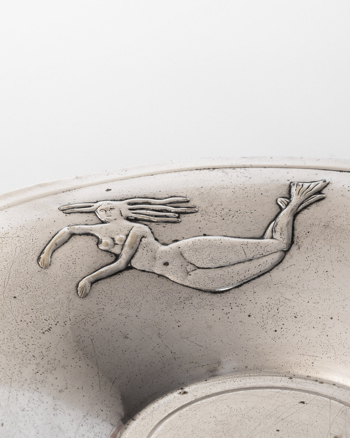 A close-up of the Decorative Bowl with Mermaids, 1930s by Spigel showcases a silver-plated surface embossed with a design of a mermaid gracefully swimming, her hair flowing elegantly. The smooth, reflective finish and slightly worn look suggest its charming Art Deco period origins.