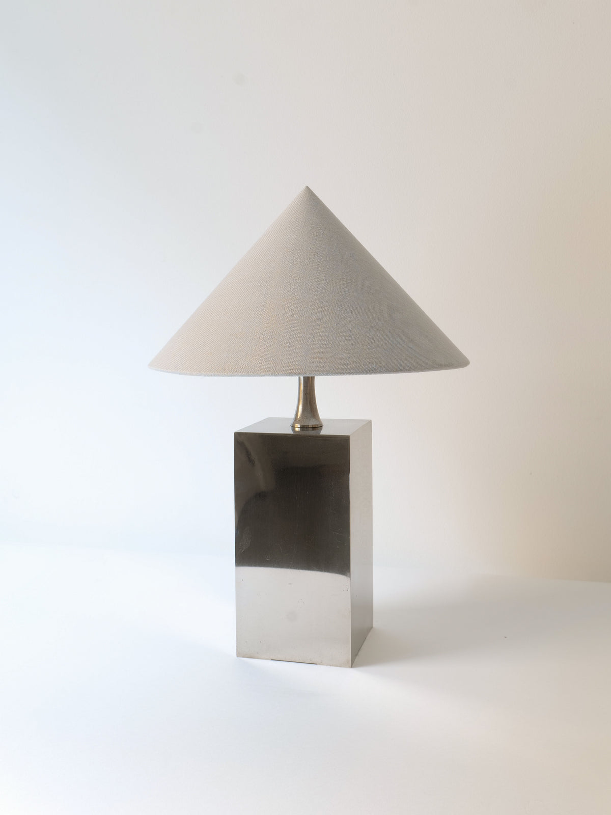 The "Barbier Small Box Lamp 1970s" by Collection apart is a minimalist table lamp, featuring a reflective, rectangular chrome-over-brass base and a conical beige lampshade, elegantly displayed against a plain white background.