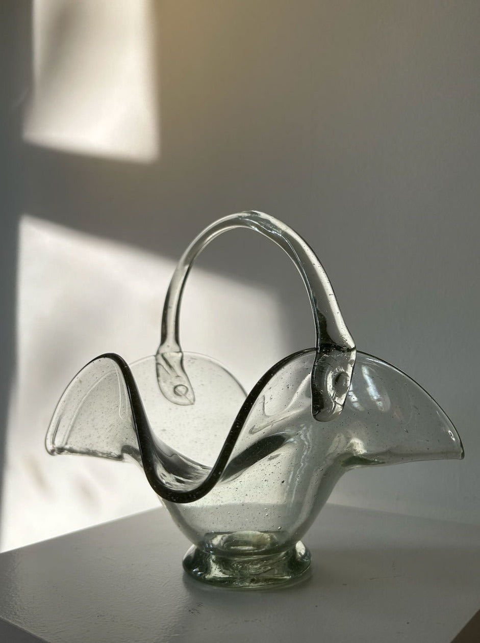 A delicate, transparent glass basket made of thick quality glass with curved edges and a handle sits on a white surface. Soft sunlight filters through a nearby window, casting shadows on the wall behind and highlighting the basket's smooth, reflective texture—reminiscent of a Les Objets Amsterdam 70's Vintage Glass Basket.