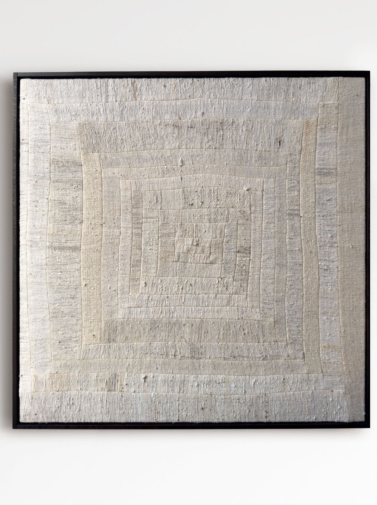 The 'To Hold', 2025 by Bec Kirby, is a textured beige textile art piece with a log cabin patchwork style. It features an intricate concentric square pattern in subtle tonal variations, framed in sleek black for a rustic and natural appearance.