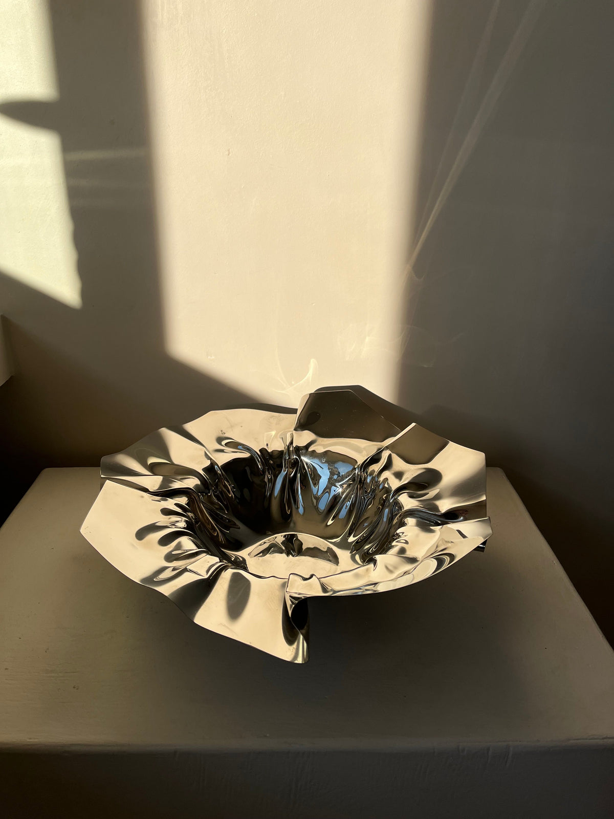 The Folded Chrome Bowl by septembre studios sits elegantly on a white surface, its crumpled, metallic appearance creating a striking folded shape. Sunlight casts dramatic shadows, emphasizing the bowl's shiny texture against the plain wall and adding a modern touch to the interior.