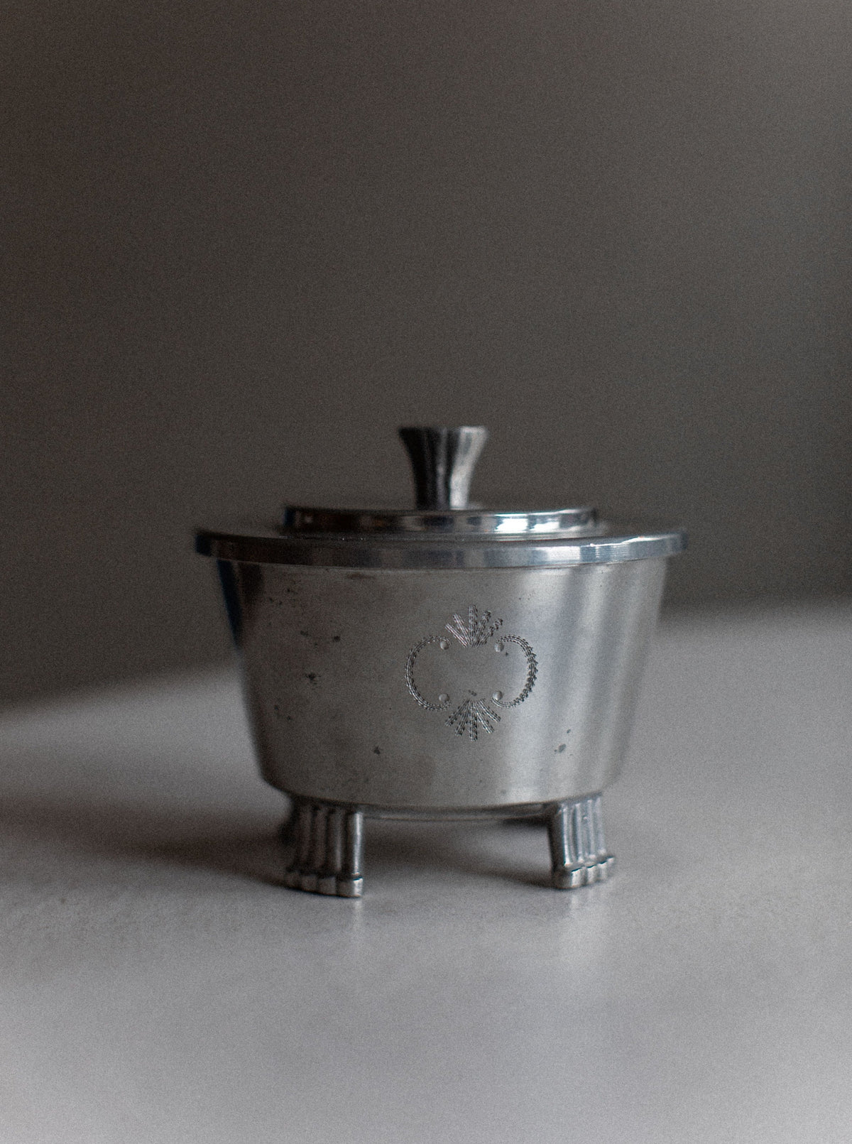 A small, silver metal container with a lid, standing on three short, leg-like supports. The container has a simple engraved design on its front, exuding minimalist charm. The background is blurred, creating focus on the vintage Art Deco Swedish Grace Tea-set by Out For Lunch.