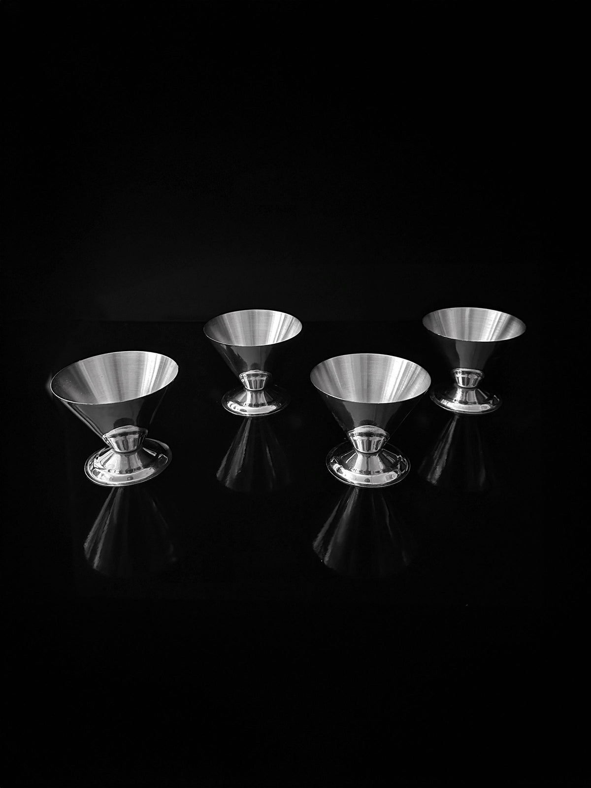 A set of four Italian Stainless Steel Dessert Cups by Dodo Vintage is arranged symmetrically on a glossy black surface, casting subtle reflections. The setup exudes minimalist elegance with a striking contrast between the metallic shine and the dark background.