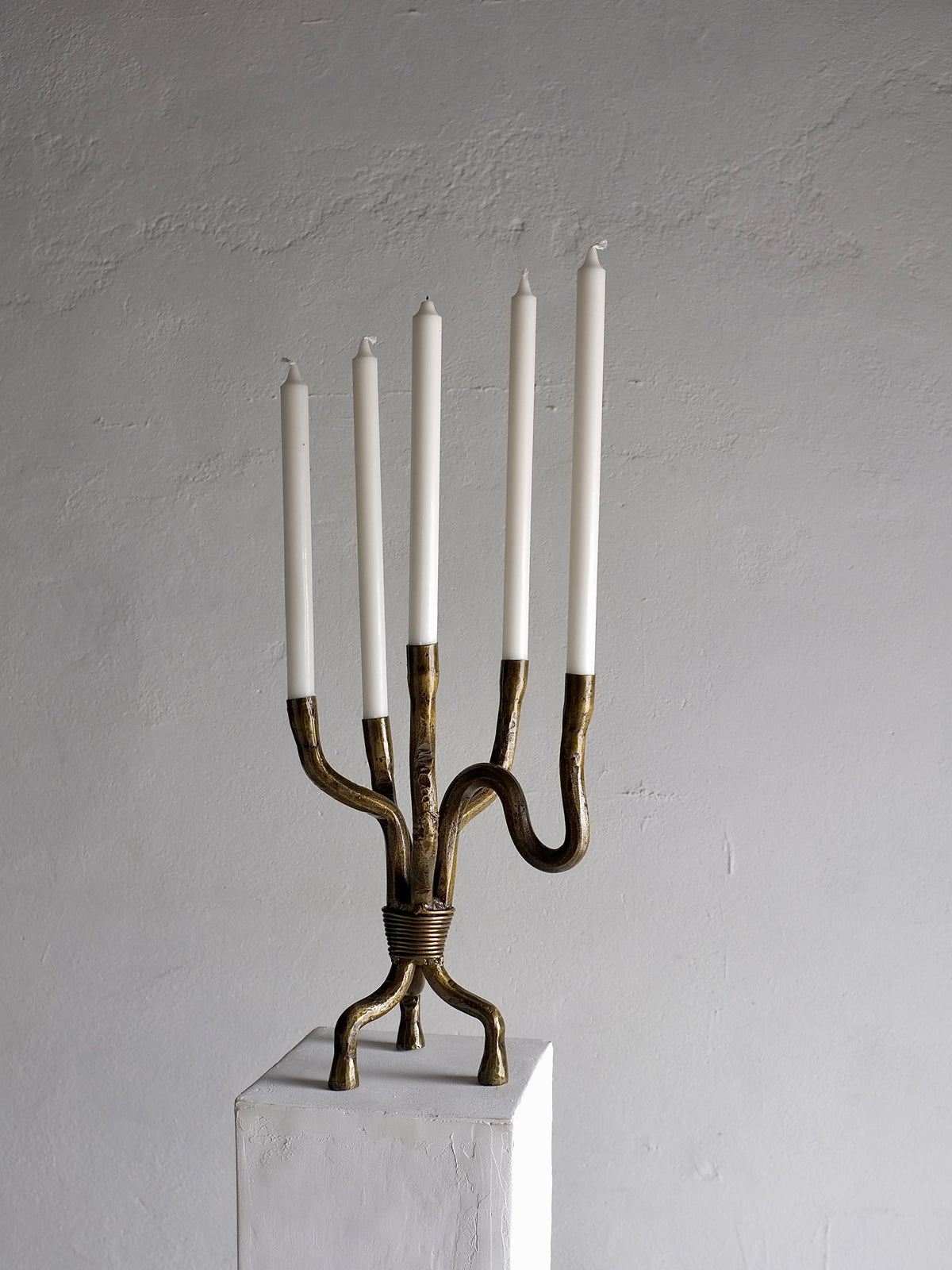 A Brutalist Brass Candelabra from the 1970s by Veter Vintage, featuring five slender, unlit white candles on a brass structure with curved and intertwined arms, stands on a white pedestal against a plain, textured grey background. This elegant piece, hailing from the Netherlands, showcases an intricate design in vintage condition.