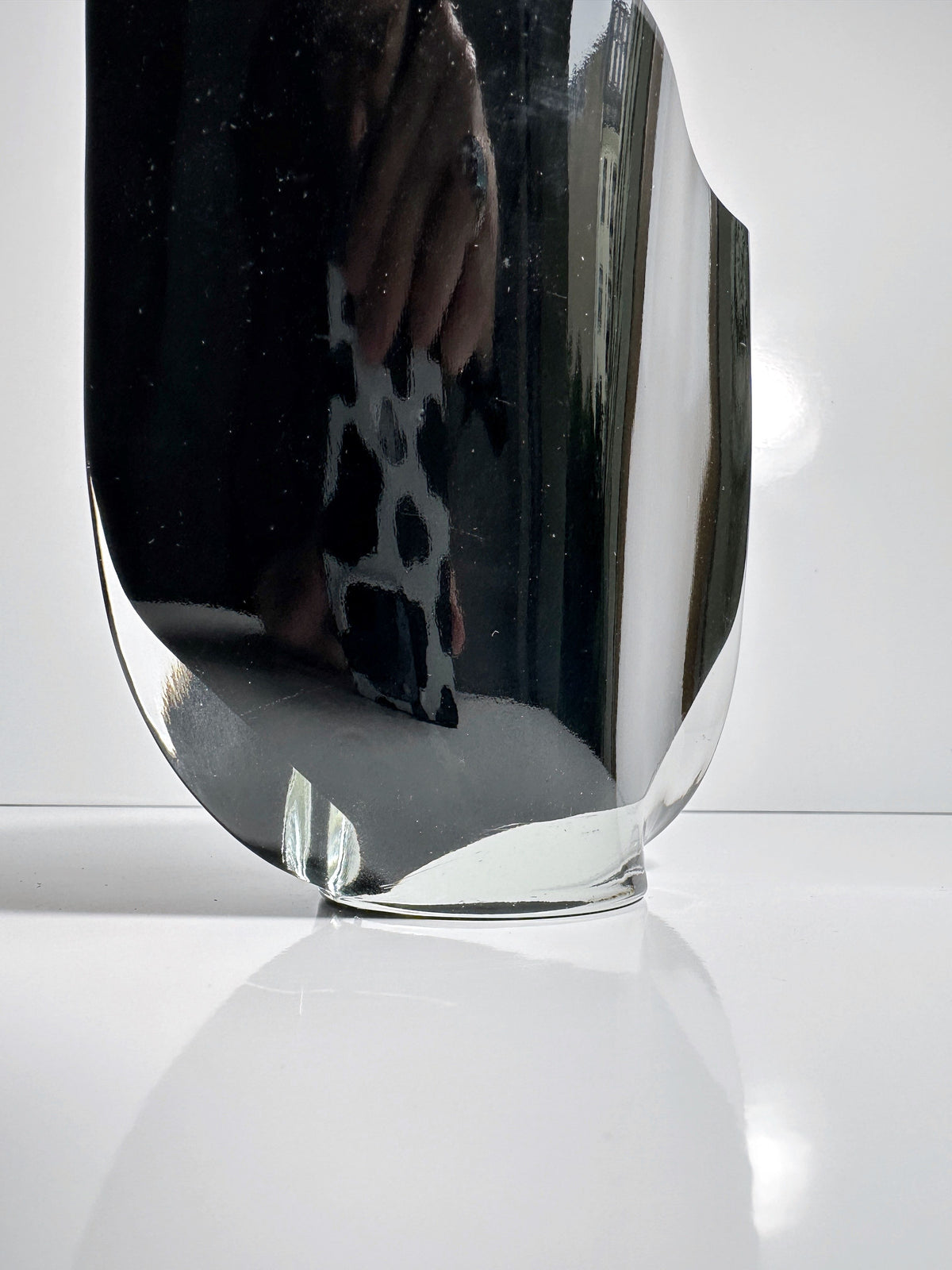A close-up photograph of a reflective Black Glass Vase by Kaj Franck from Dodo Vintage, resting on a glossy white surface. The smooth, curved surfaces of the vase reflect its surroundings, including a hand holding a phone with a patterned case. The background is out of focus.