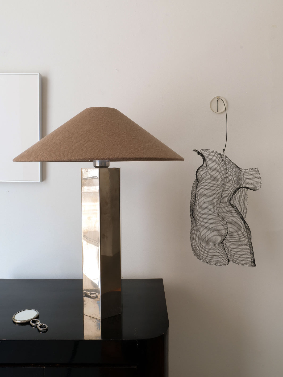 The Grand Hexagonal Lamp by Collection apart, featuring a beige shade, sits on a dark table beside glasses and keys. On the wall are a wire mesh torso sculpture and white-framed artwork.