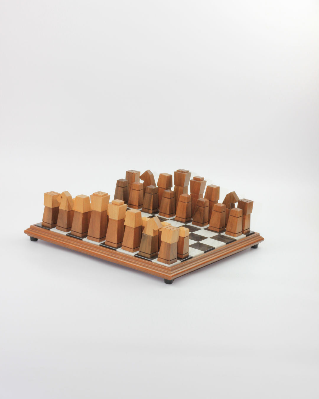 A minimalist Boga Avante Shop Wooden and Marble Chessboard is displayed on a simple wooden board. Each chess piece is geometrically shaped with varying heights and forms, all in natural wood tones, arranged neatly in their starting positions for a game. The background is plain white.