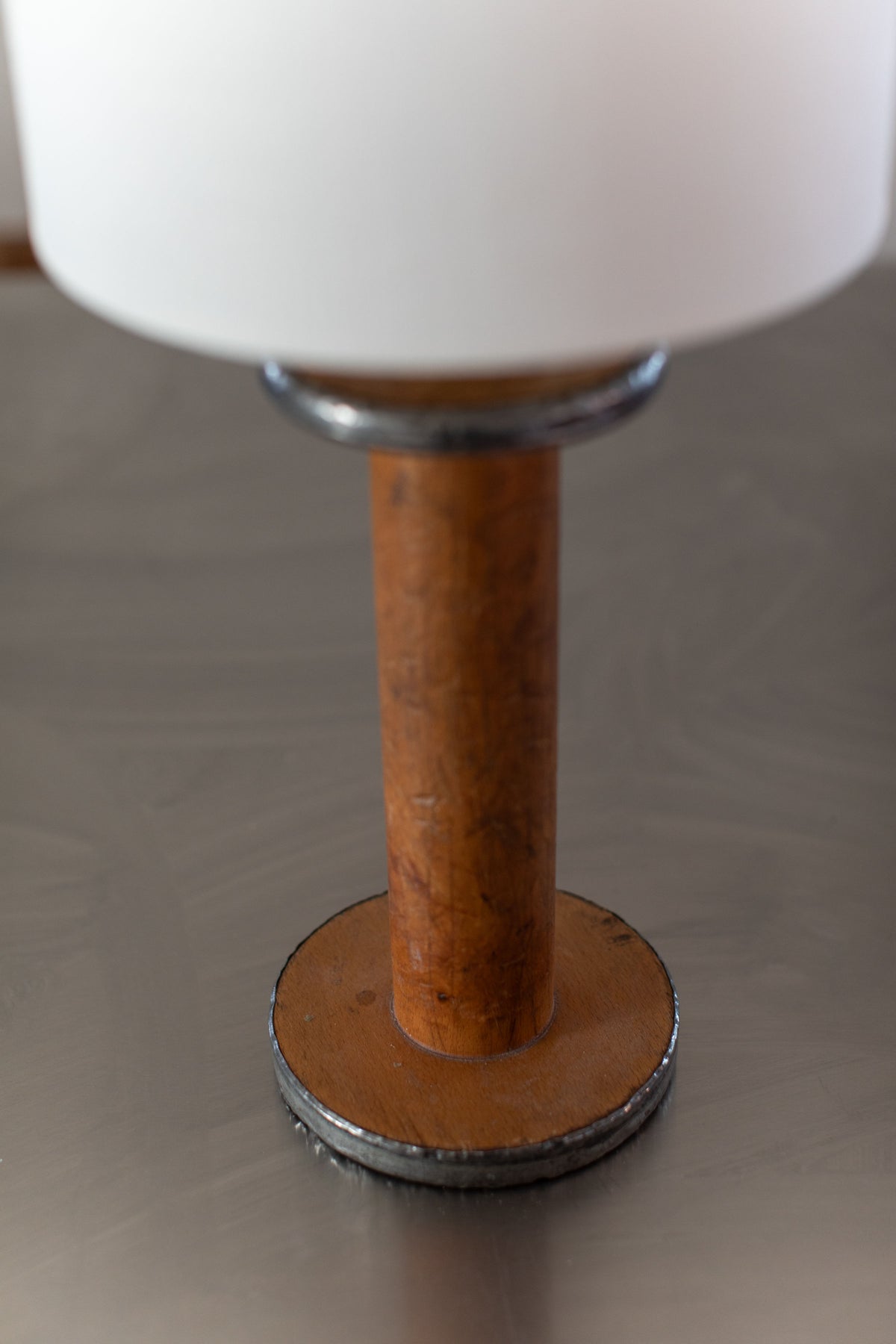 A close-up image of the Large Mid-Century Lamp by Out For Lunch, featuring a wooden handle base and a round metal base. The lamp is topped with a white lampshade, and the surface appears to be worn with some visible scratches. The background is a smooth, reflective surface.