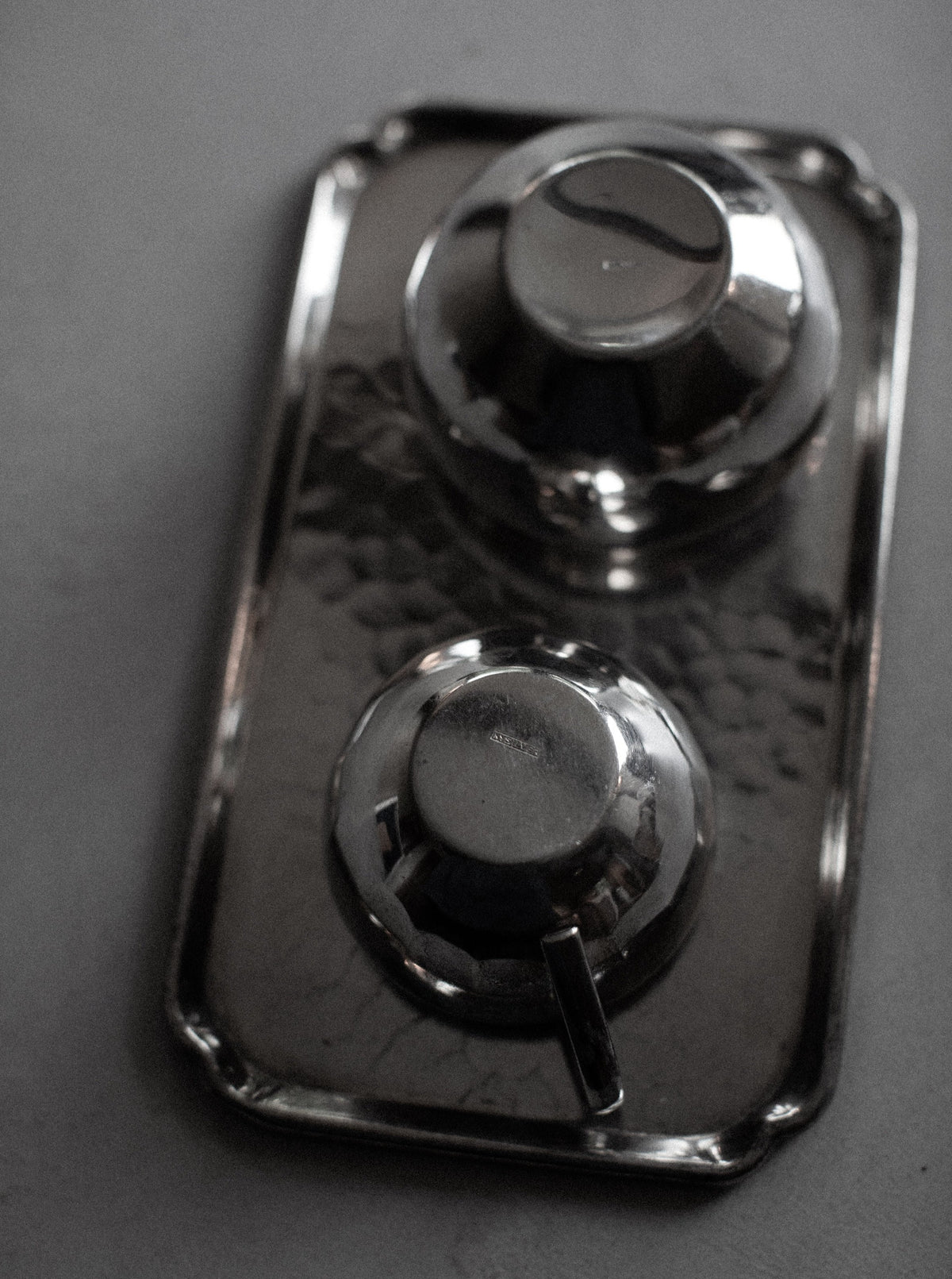 A rectangular, hammered metal tray holds two rounded, dome-shaped containers with lids and small handles. The Out For Lunch Art Deco Creamer & Sugar Bowl reflect light and create a slightly blurred, muted appearance in the grayscale image, exuding an air of vintage glamour.
