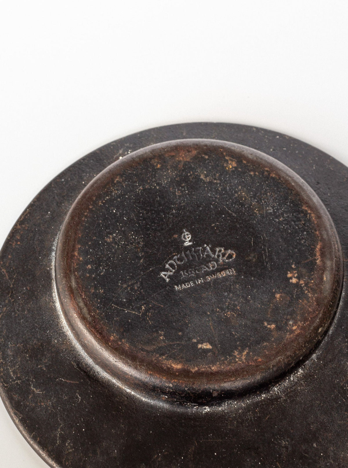 The image shows the underside of a circular, worn-out, dark-colored object that resembles an Art Deco Zodiac Signs Decorative Dish by Spigel. The bottom features a small raised ring and is inscribed with the words "ADDITARD" and "MADE IN SWEDEN." The surface appears aged and scratched, reflecting its Scandinavian vintage design.