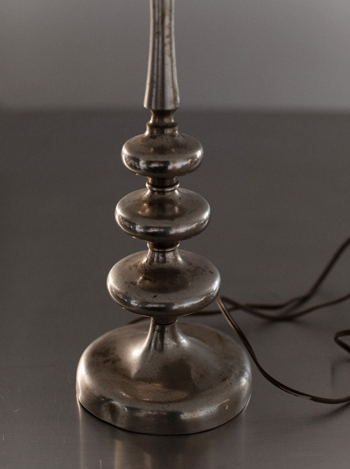 A close-up of the 1930's Table Lamp by Out For Lunch features a metallic base with a round, tiered design. The base exudes vintage decor appeal with slight tarnishing and is placed on a reflective surface. The partially visible electrical cord extending from the base enhances its Art Deco charm.