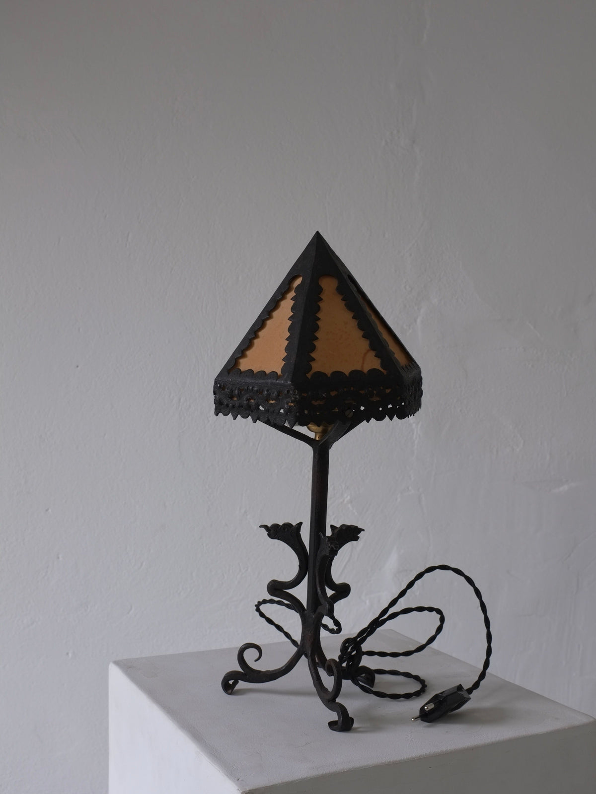 The Veter Vintage Wrought Iron Dragon Table Lamp, Italy 1910s, features an ornate black base and a small beige pyramid shade. It sits on a white pedestal against a light gray wall, with its cord coiled neatly beside it.
